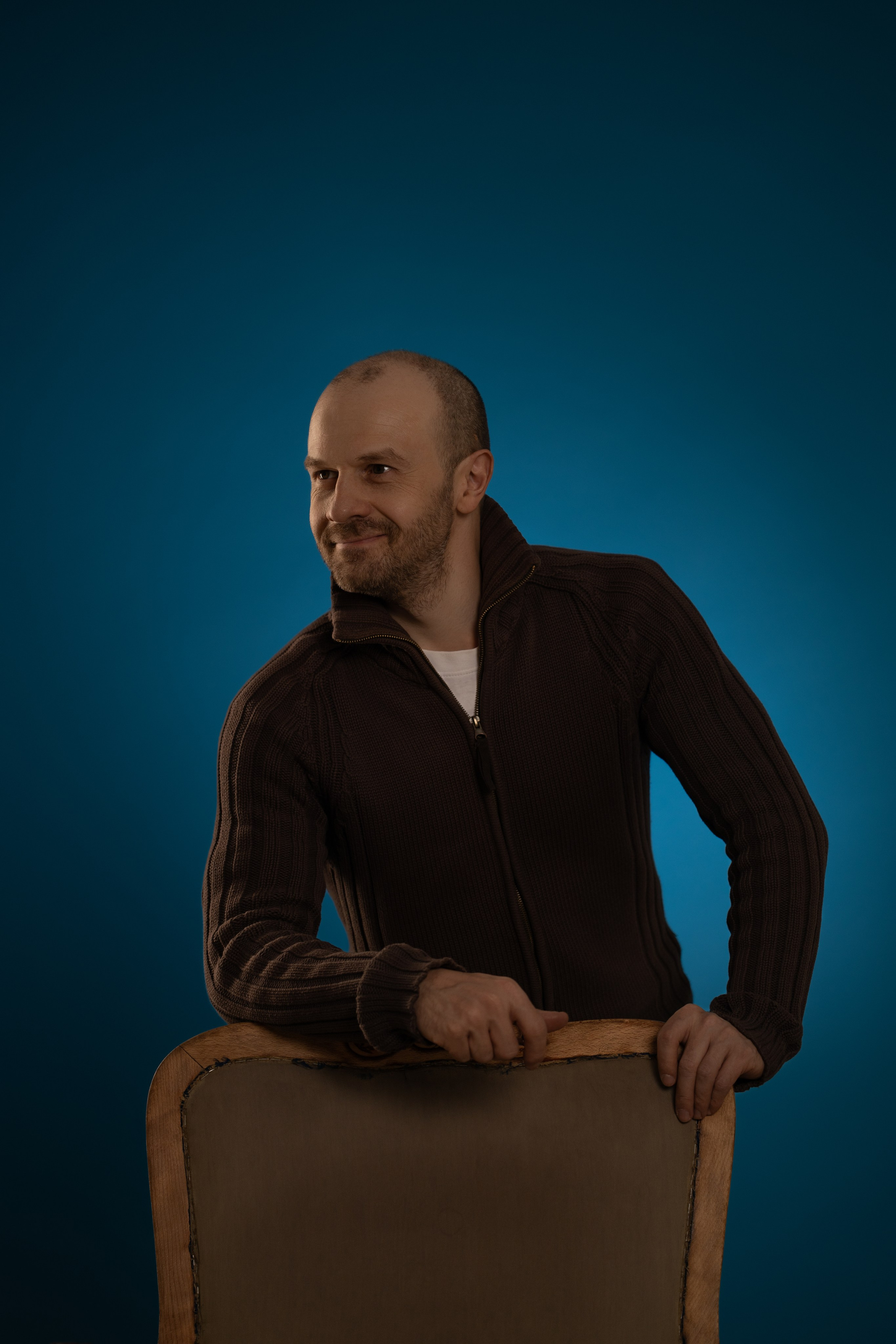 Портрет мужчины в коричневом свитере, облокотившегося на кресло, на синем фоне.