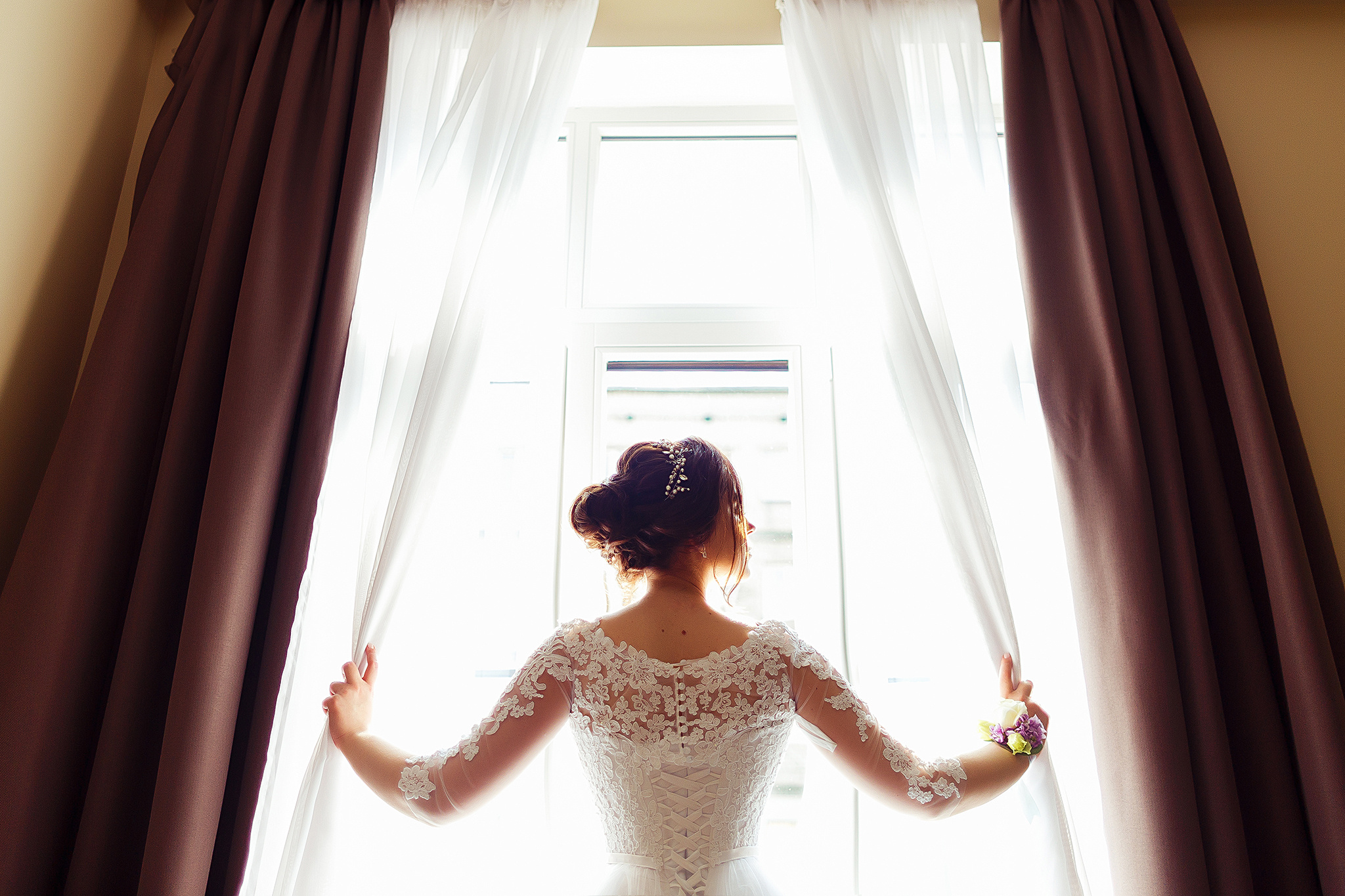 Невеста в легком платье трогает шторы в стильном номере отеля