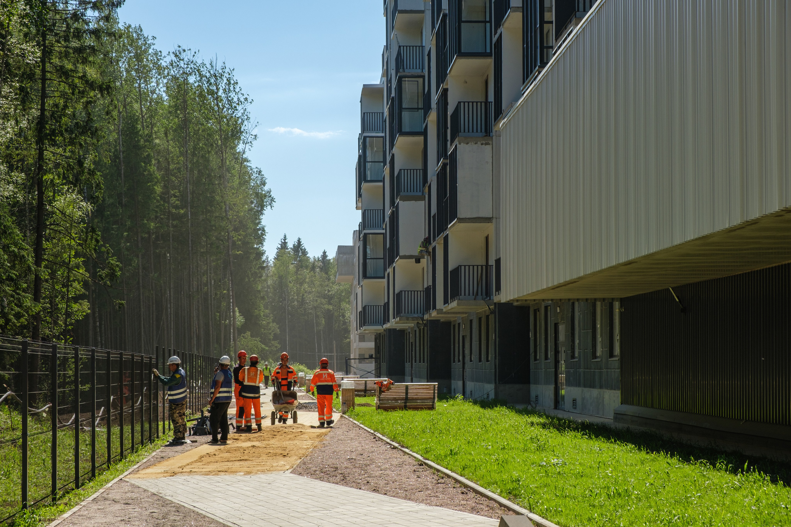 Рабочие в защитной одежде занимаются благоустройством территории, возможно, они укладывают тротуарную плитку. Рядом с ними расположены строительные материалы и инструменты.
