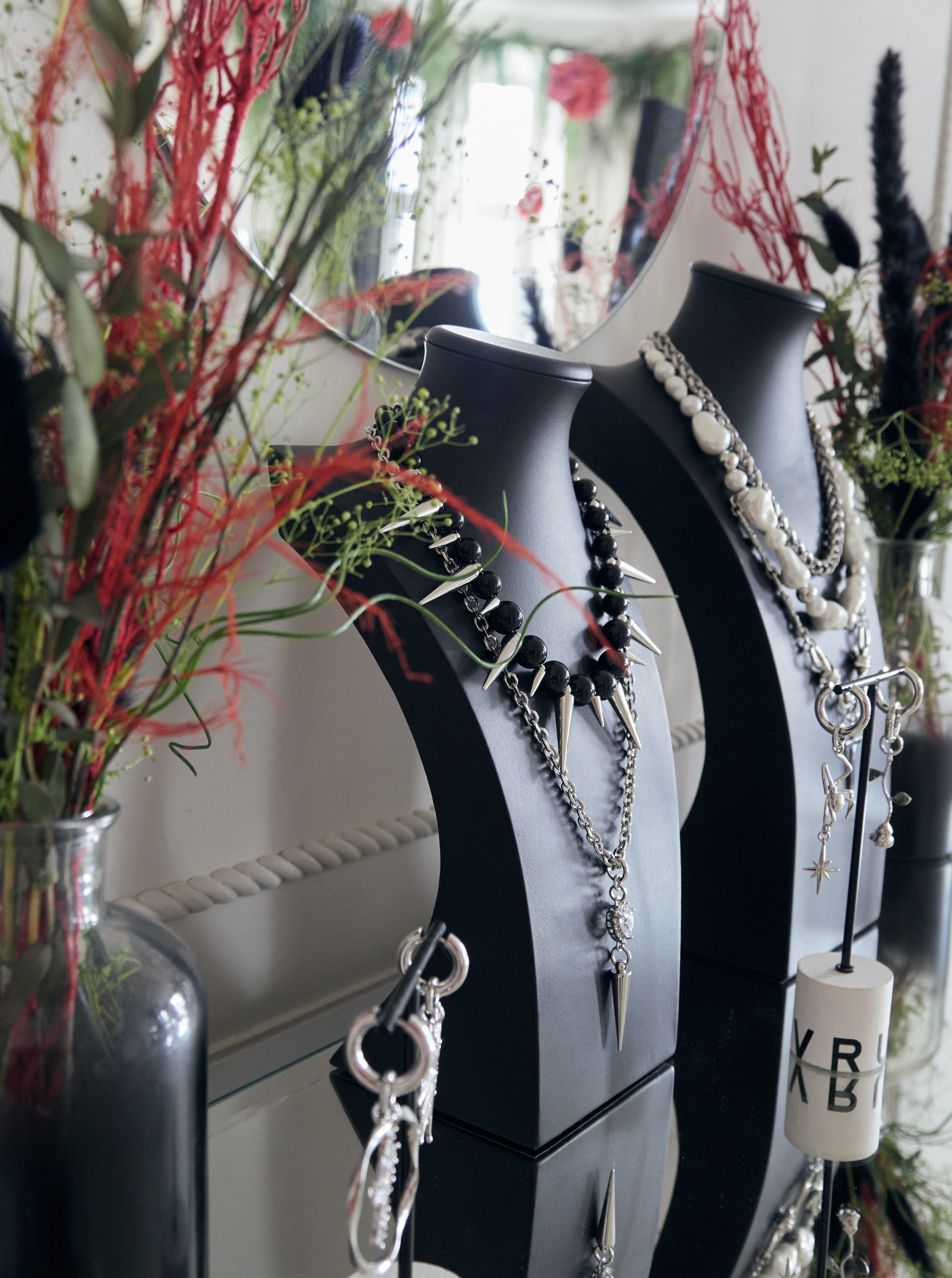 Выставка ювелирных изделий на зеркальной поверхности: два черных бюста на шее и многослойные ожерелья черного, белого и серебряного цветов. Вокруг витрины расположены вазы с листвой и декоративными элементами, создающие стильную и элегантную презентацию.