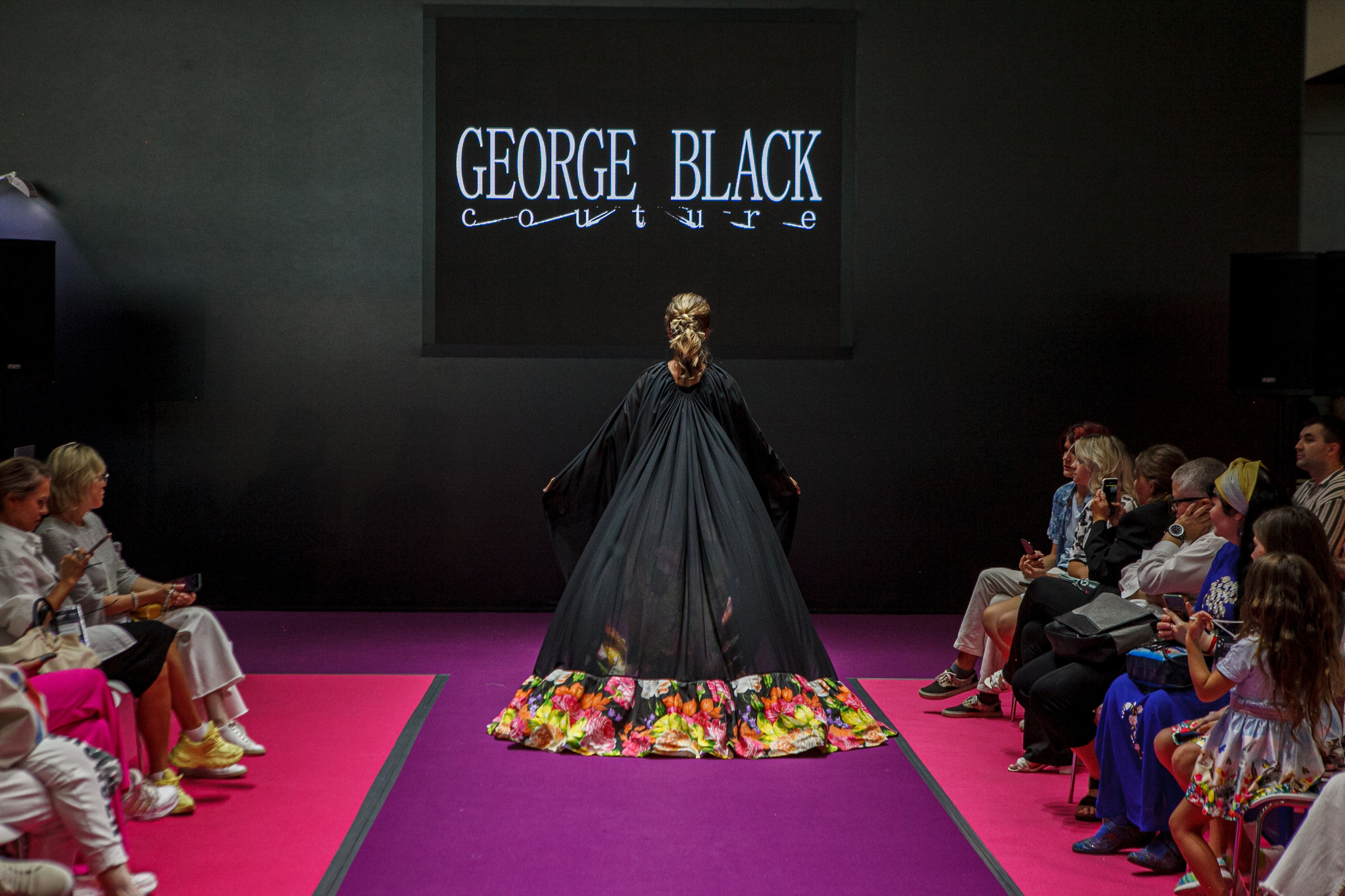  "Модели в ярких нарядах проходят по подиуму во время показа мод, освещенные яркими софитами и окруженные восторженными зрителями."