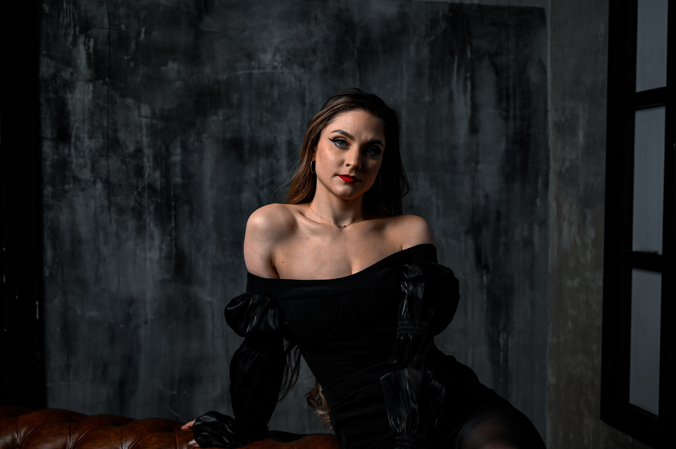 фотосессия девушки, которая сидит на кожаном диване в темной комнате. Она одета в черное платье с открытыми плечами, которое подчеркивает её фигуру и создает элегантный образ. Её длинные волосы свободно падают по плечам, а взгляд направлен прямо в камеру, демонстрируя уверенность и сосредоточенность. На лице у нее акцентный макияж с ярко-красными губами, который контрастирует с тёмным фоном. Вокруг модели видны элементы интерьера, такие как окно со ставнями и текстурная стена, что добавляют атмосферу загадочности и интриги. Фотография передает чувственную и стильную атмосферу.