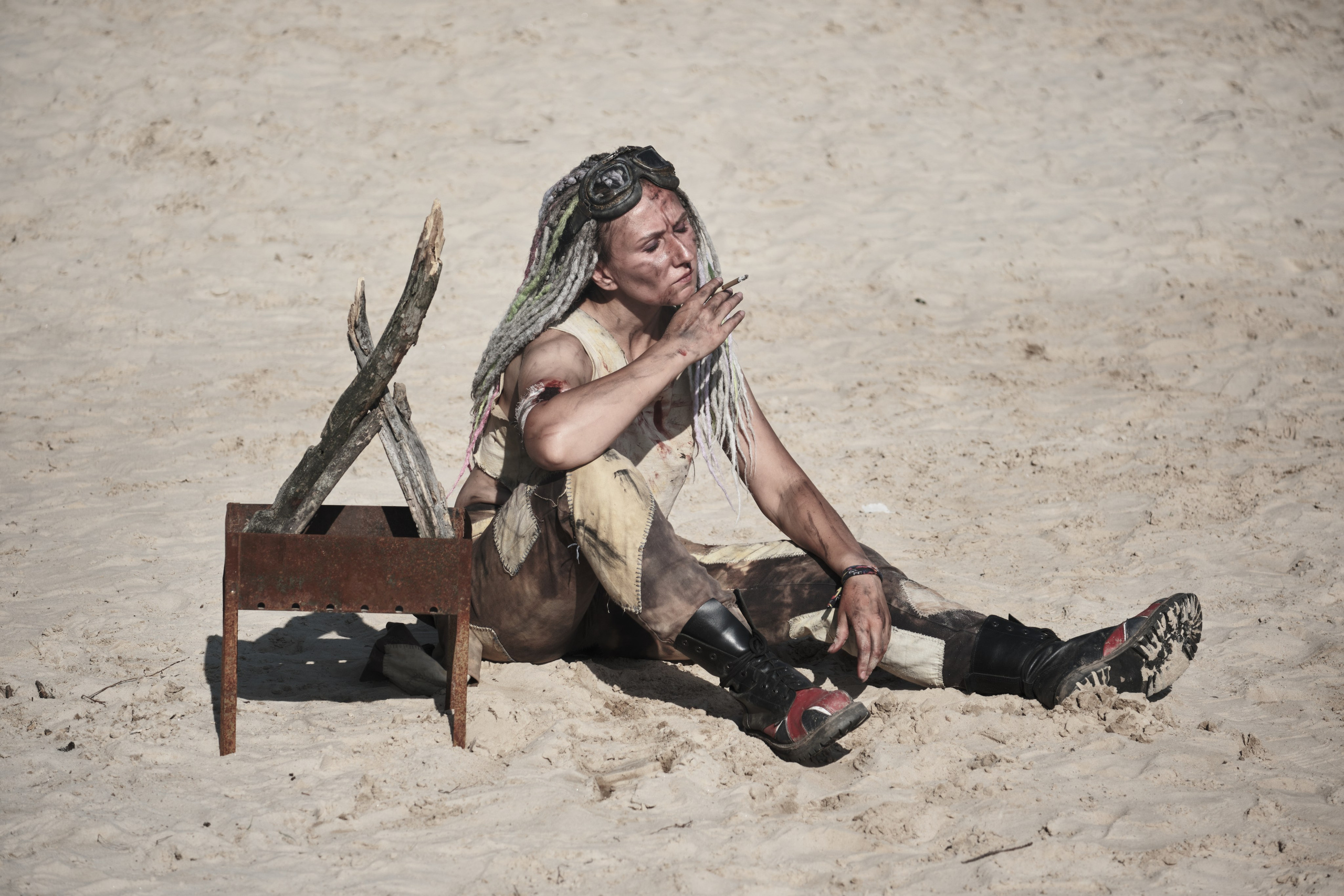 Девушка с дредами курит у мангала на песке в стиле пост апокалипсис