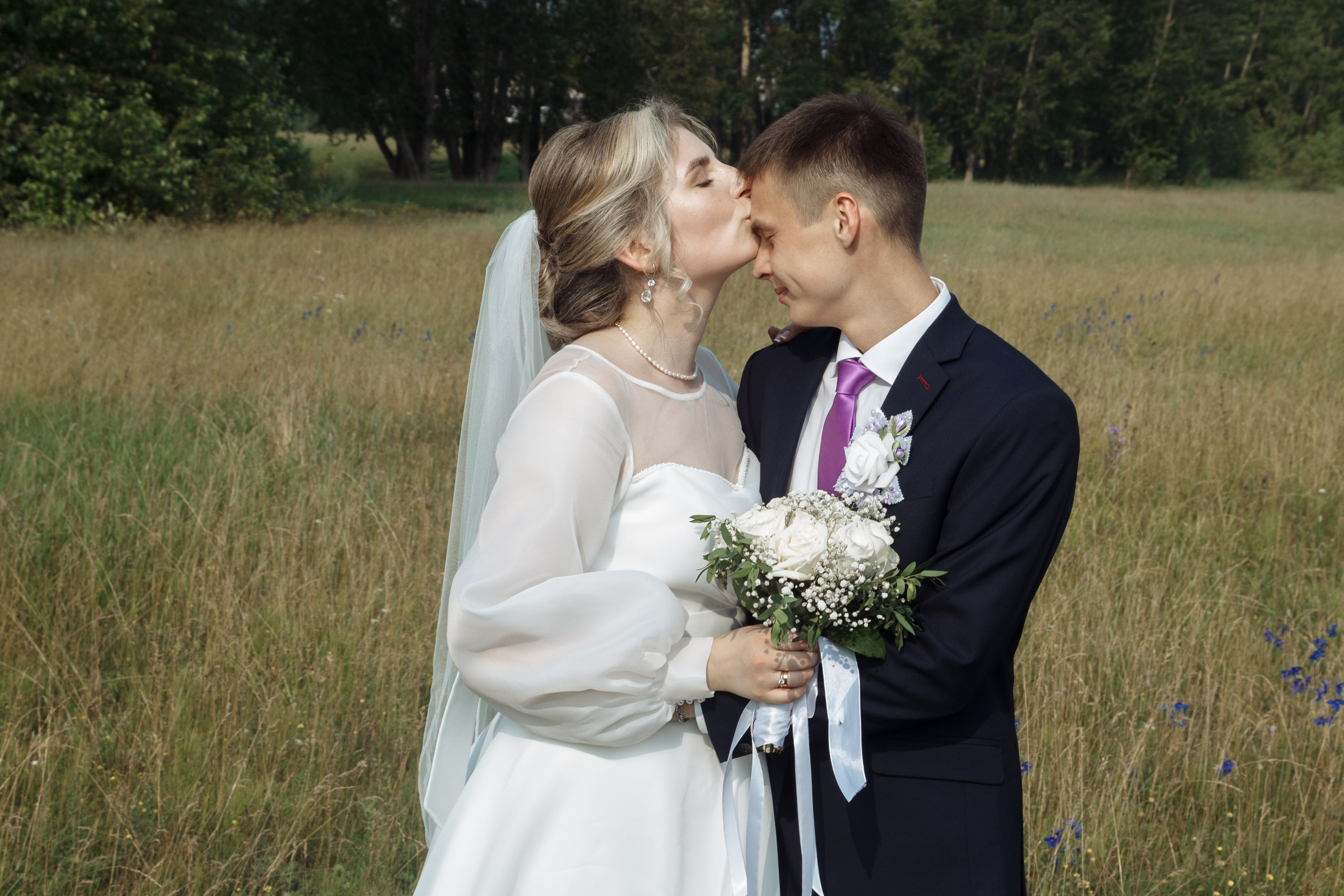 Жених целует невесту в лоб на фоне поля, невеста с букетом, момент нежности.
