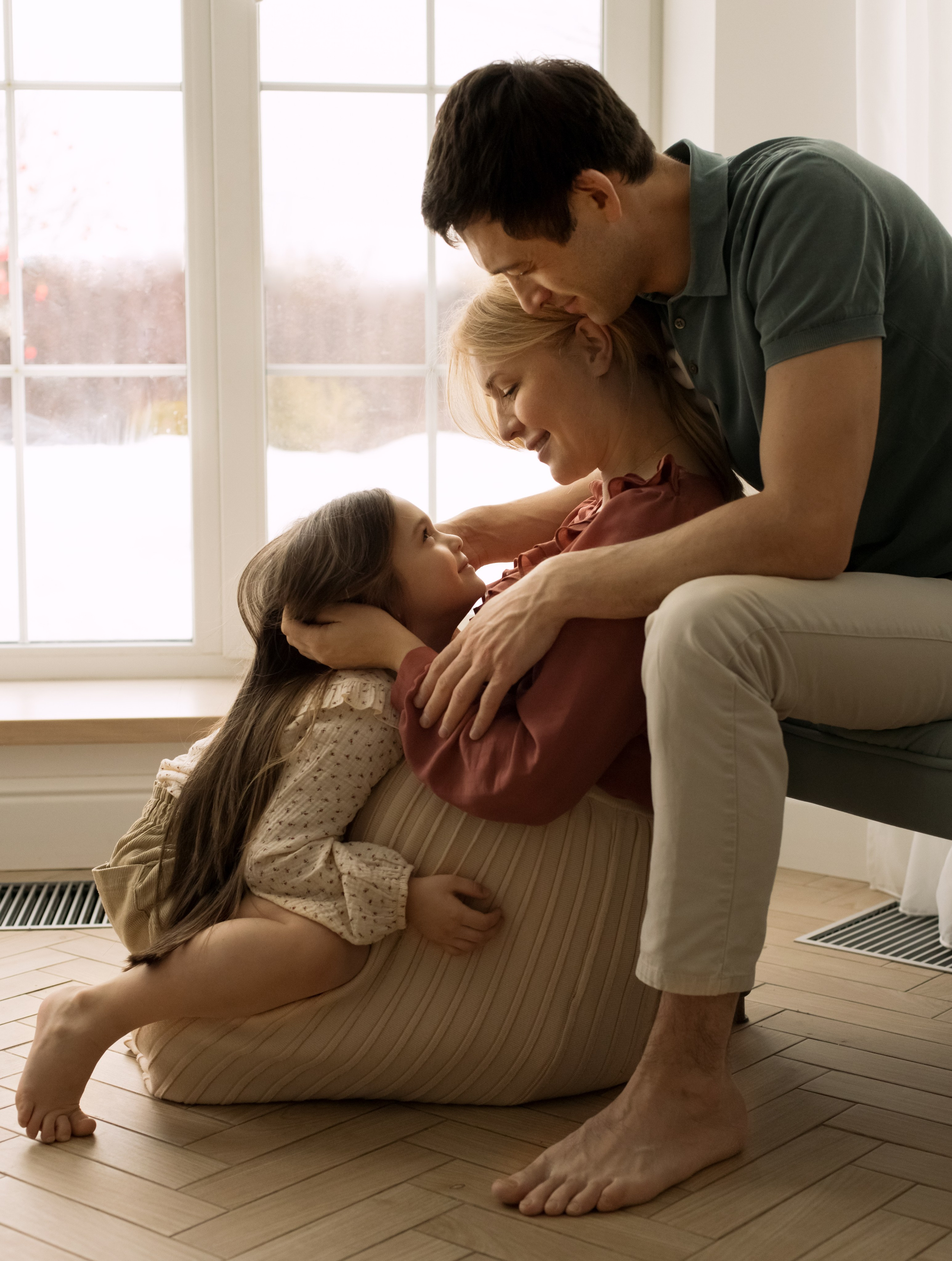 Беременная мама, папа и дочка обнимаются сидя у большого окна.