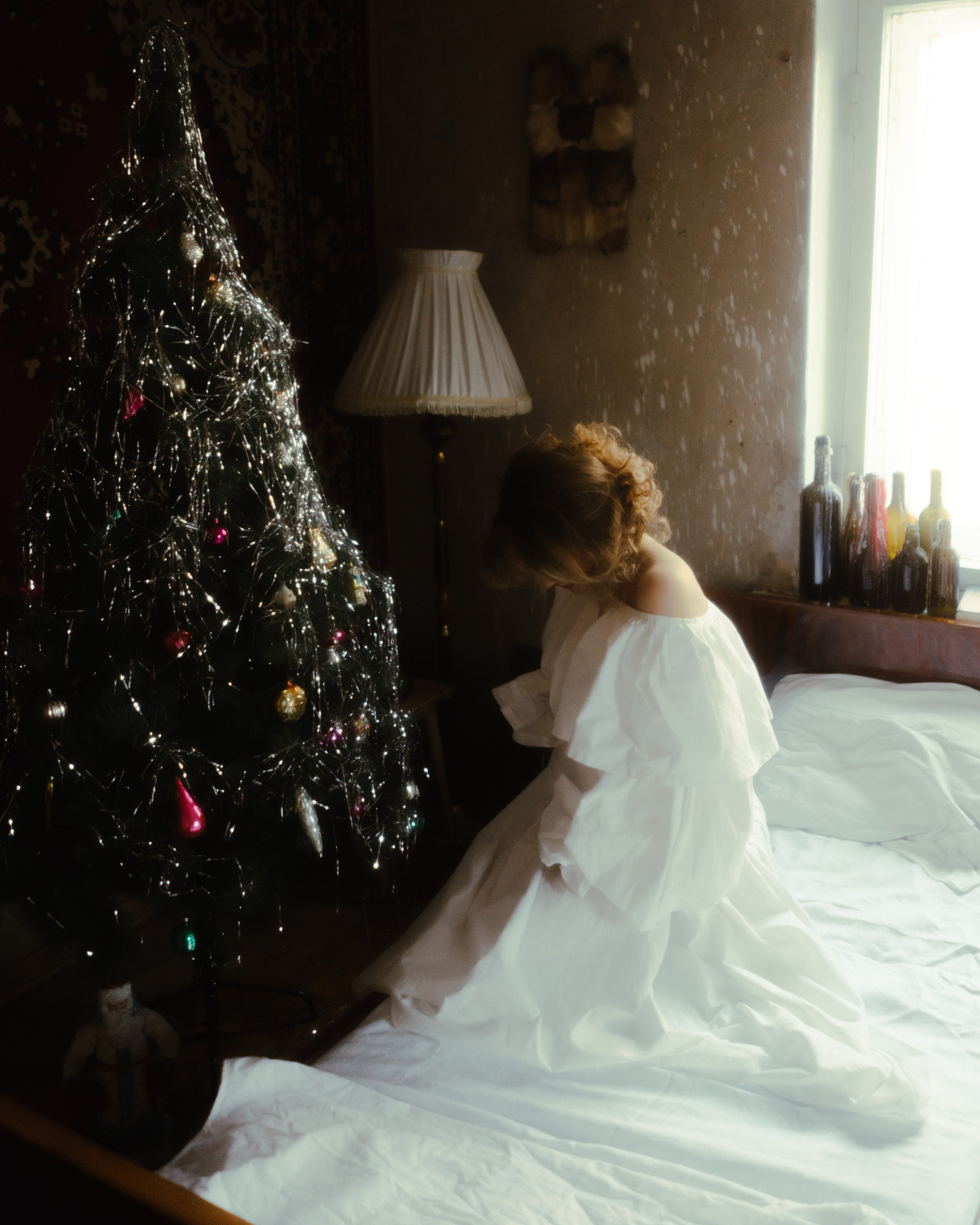 Беременная в белом платье рядом с елкой на кровати, пленочная атмосфера, мягкий свет и винтажный интерьер, студийная фотосессия беременности в СПб.