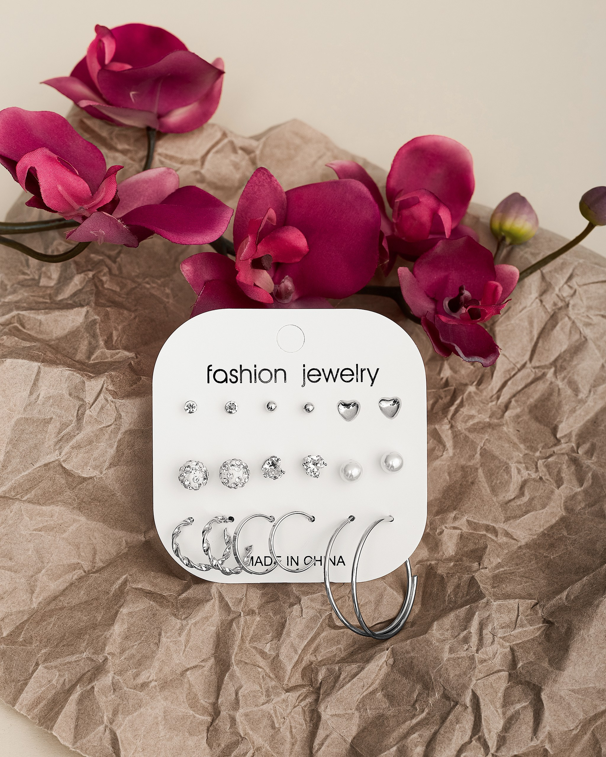 Серьги-гвоздики и кольца на подложке, рядом розовые орхидеи, бежевый фон