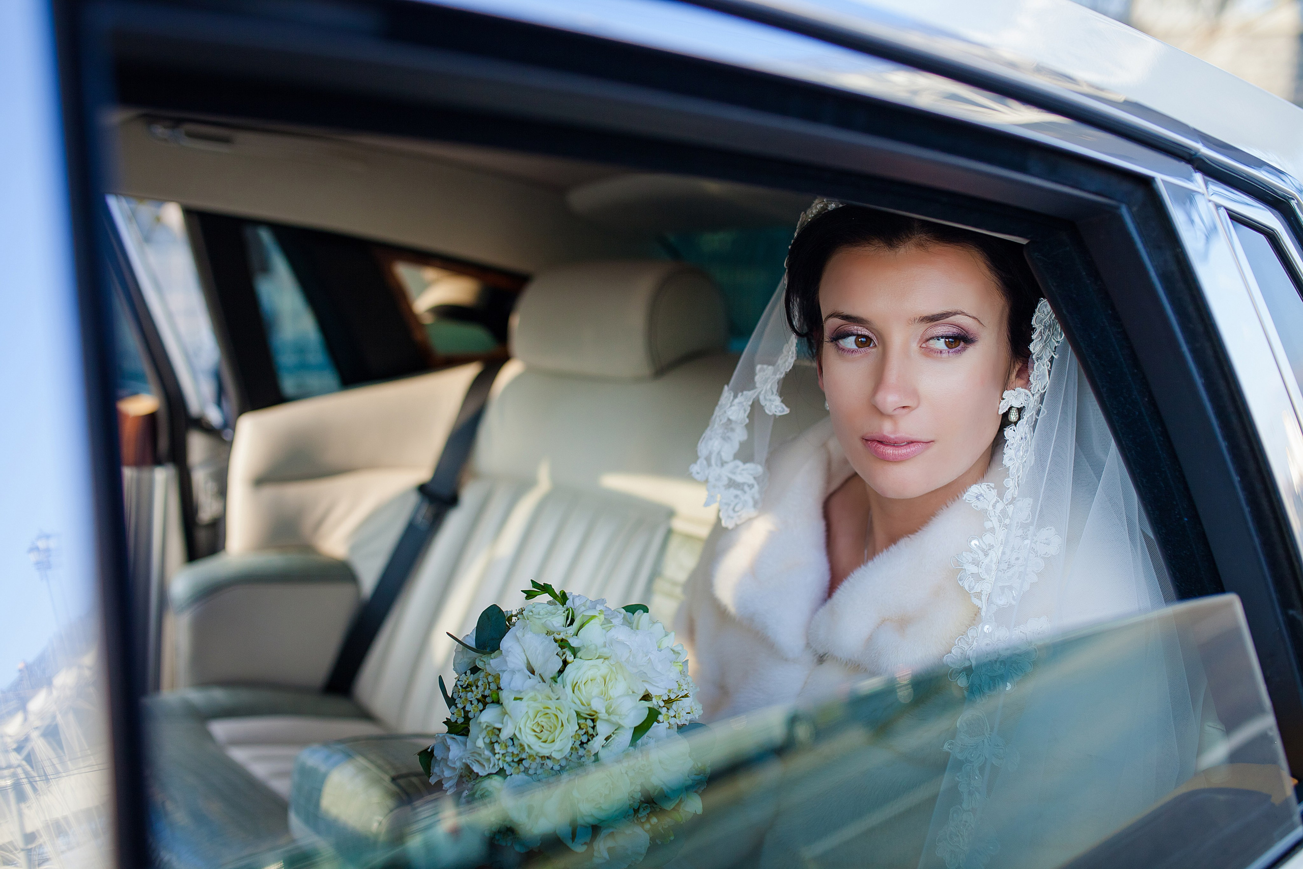 Невеста в белом платье из букетом сидит в машине и смотрит в окно