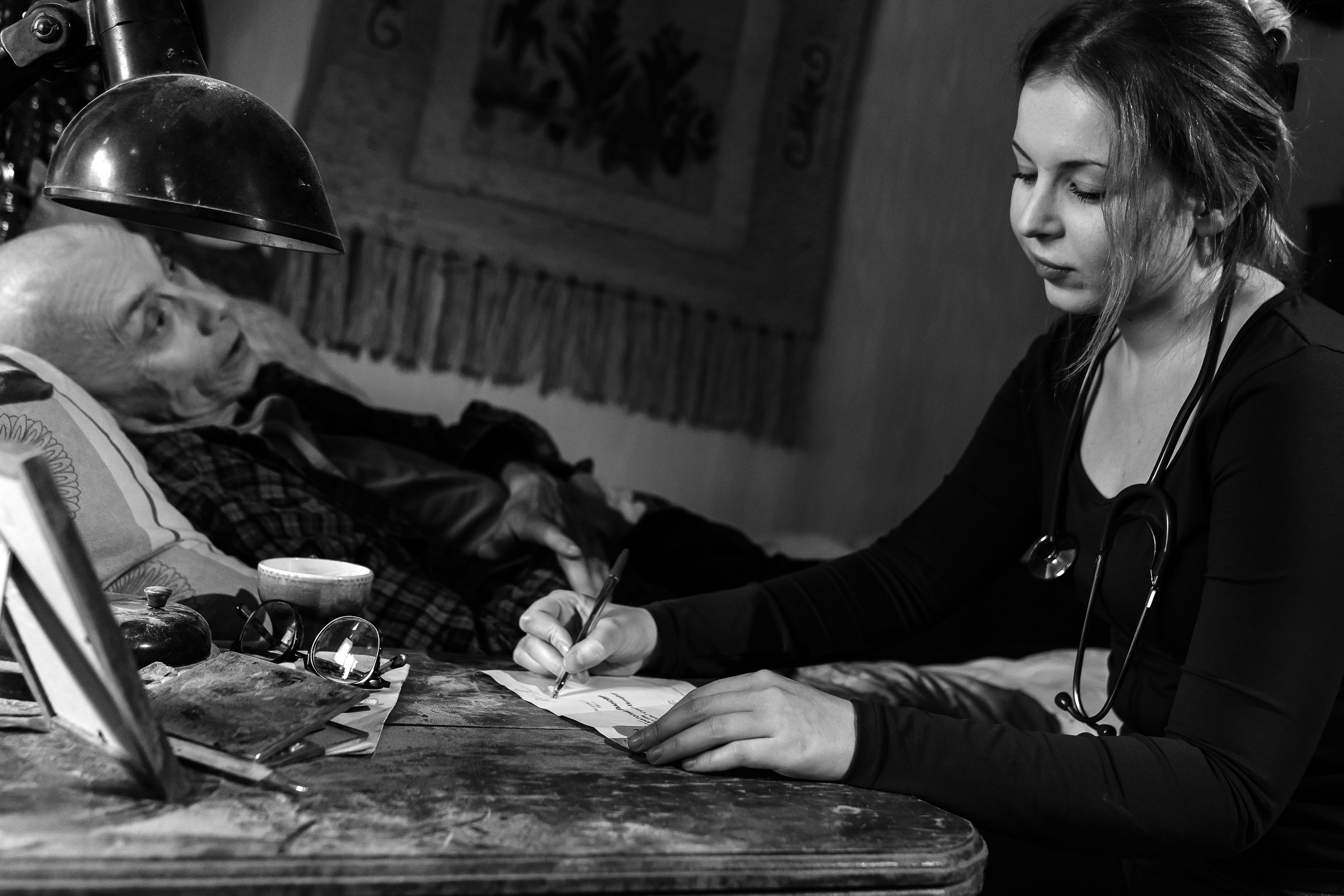 Черно-белая сцена врачебного приема на дому: врач с фонендоскопом выписывает рецепт у постели больного. Кадр из фильма «Участковый терапевт» в Петербурге. Фото Евгения Мальцева, evrednix.ru.