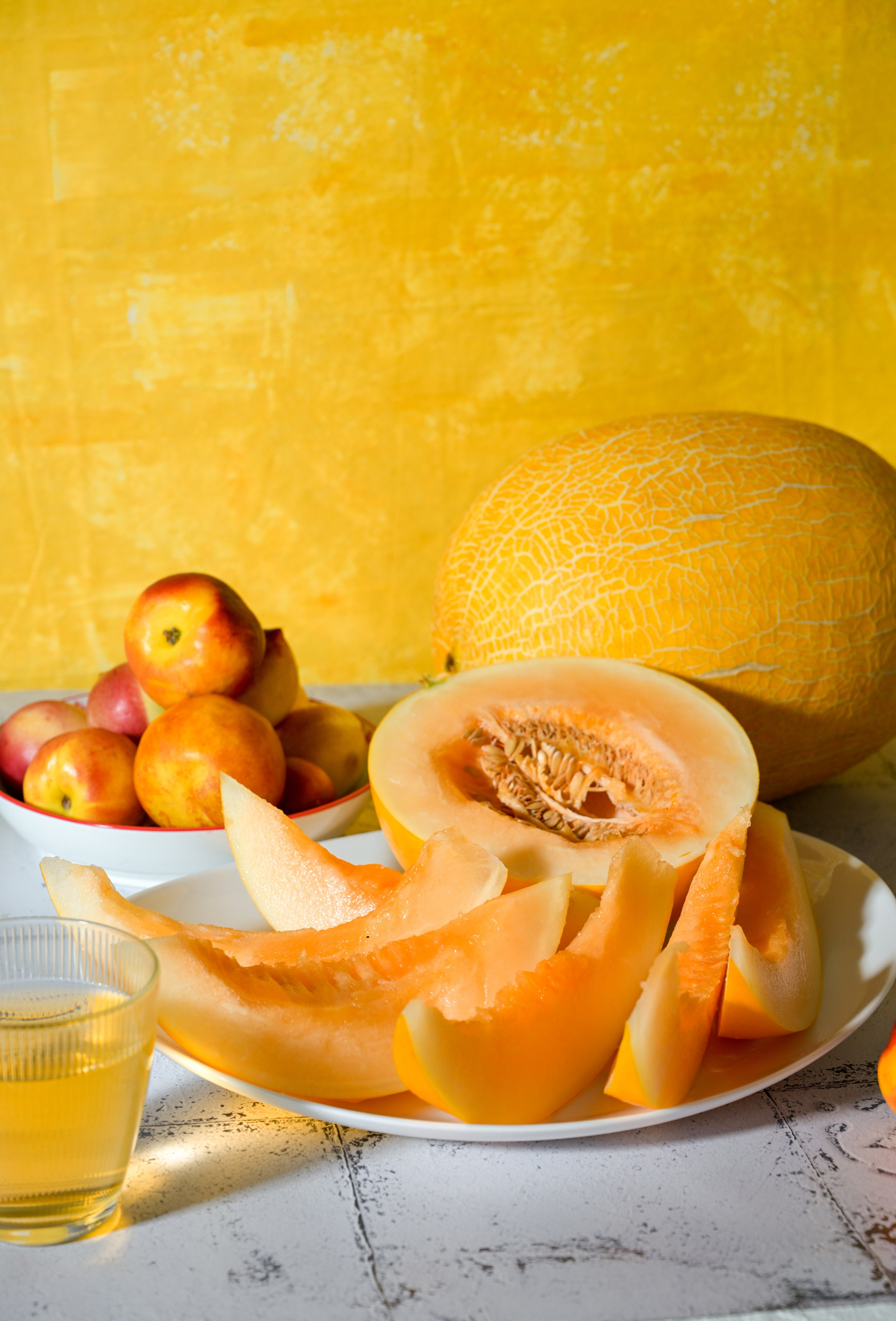 Нарезанная дыня на белой тарелке, целая дыня, нектарины в миске и стакан сока на светлом фоне с жёлтой стеной