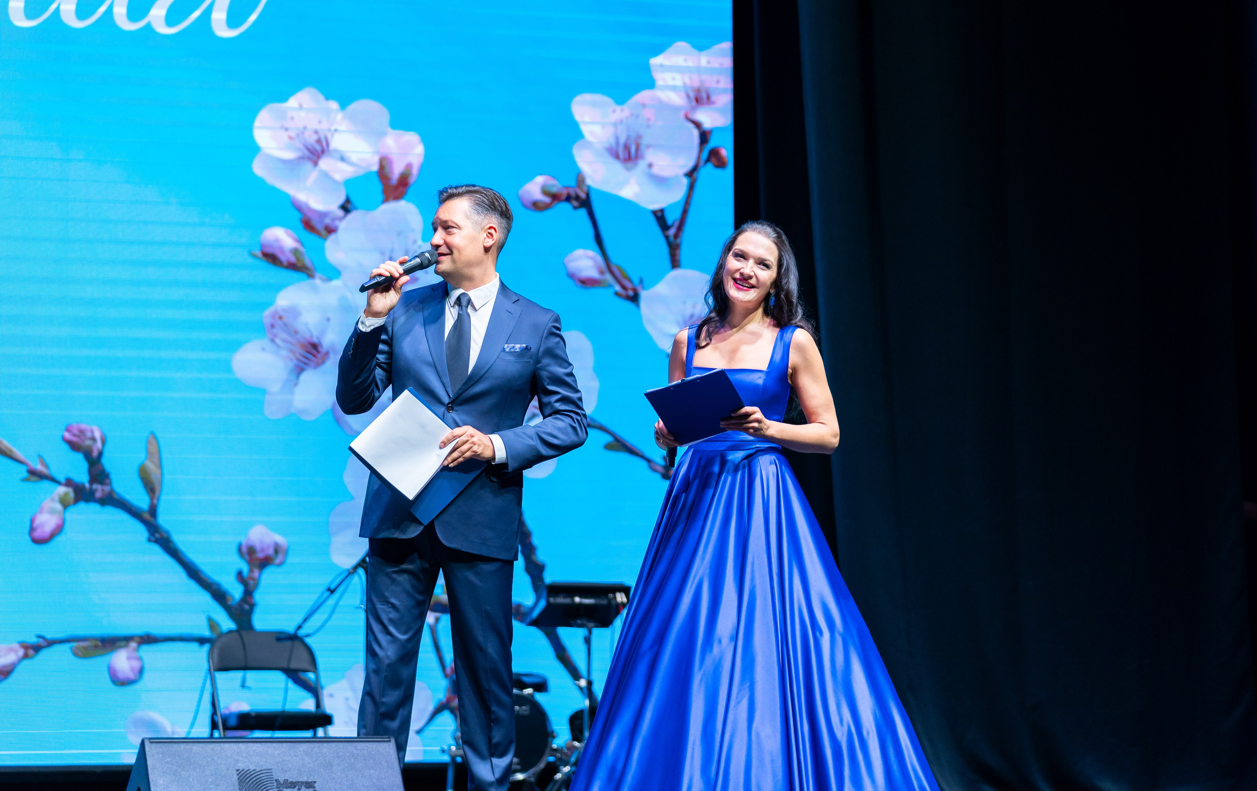 Мужчина в синем костюме с микрофоном и женщина в длинном синем платье на сцене. Ведущие церемонии награждения Дня местного самоуправления в Санкт-Петербурге | Фото: Евгений Мальцев.