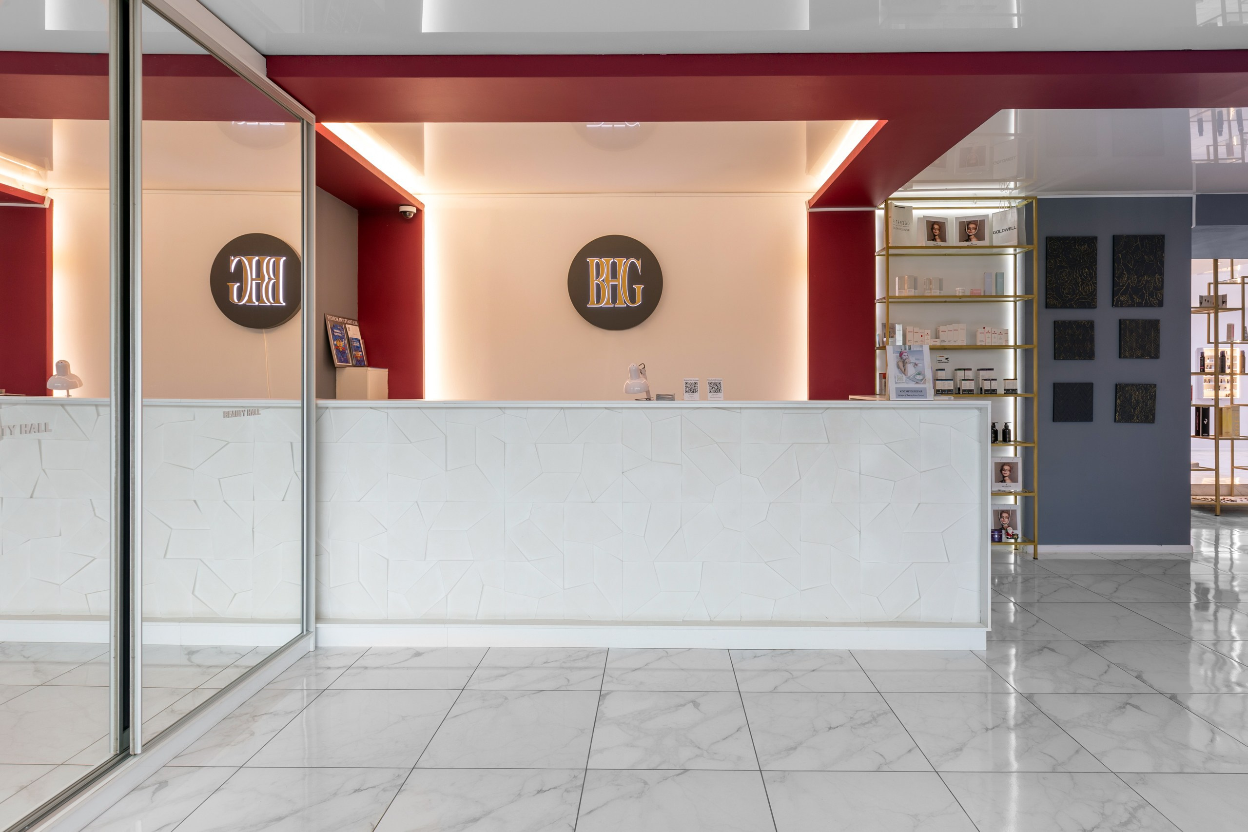 Фото интерьера салона красоты. Каменная светлая стойка администратора и большое зеркало. Красная и бежевая стена с логотипом салона и подсветкой.
