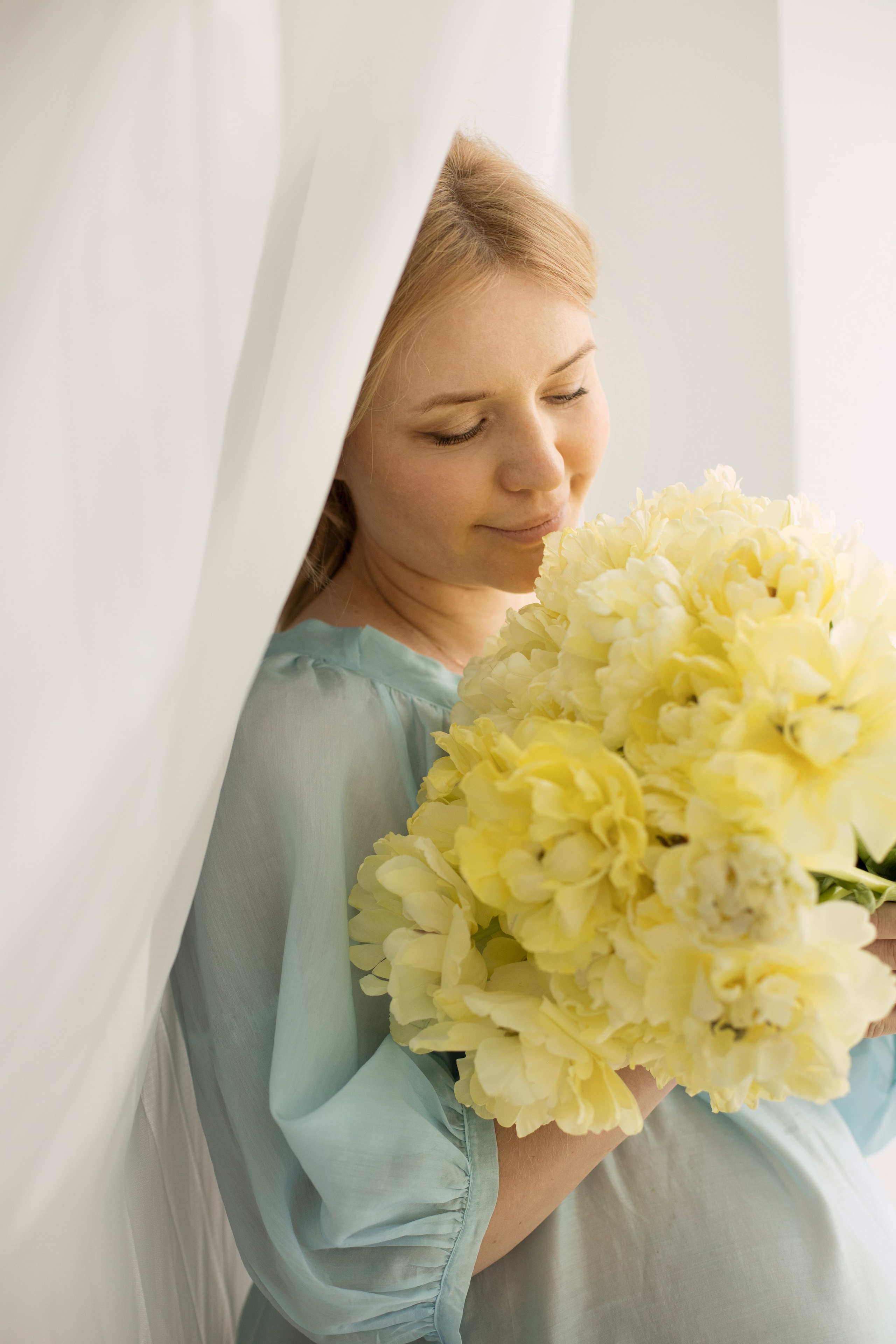 Беременная женщина в  голубом платье стоит у окна и смотрит на букет желтых пионов .