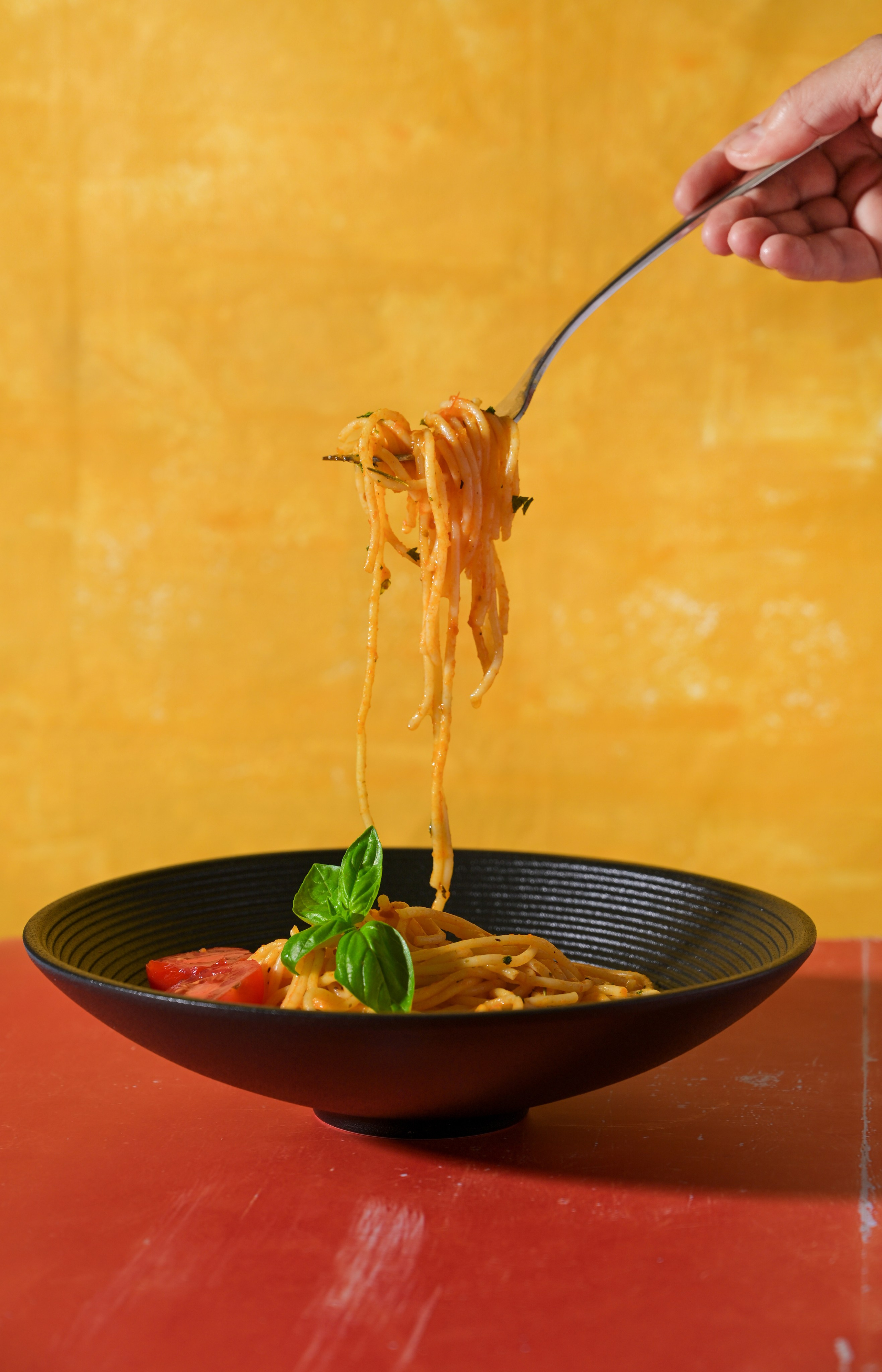Тарелка с пастой в томатном соусе с базиликом, черная посуда, рука держит вилку с макаронами на фоне желтой стены
