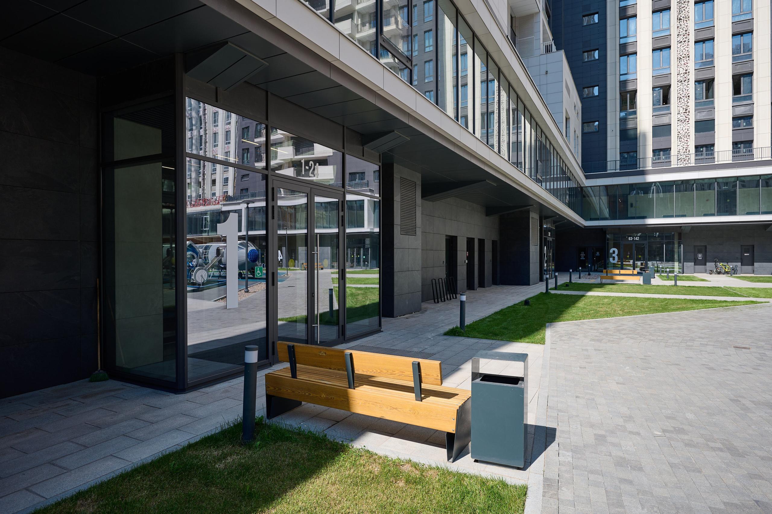 Вид на стеклянный фасад современного жилого комплекса с лавочками и мусорными баками во внутреннем дворике; яркий солнечный день