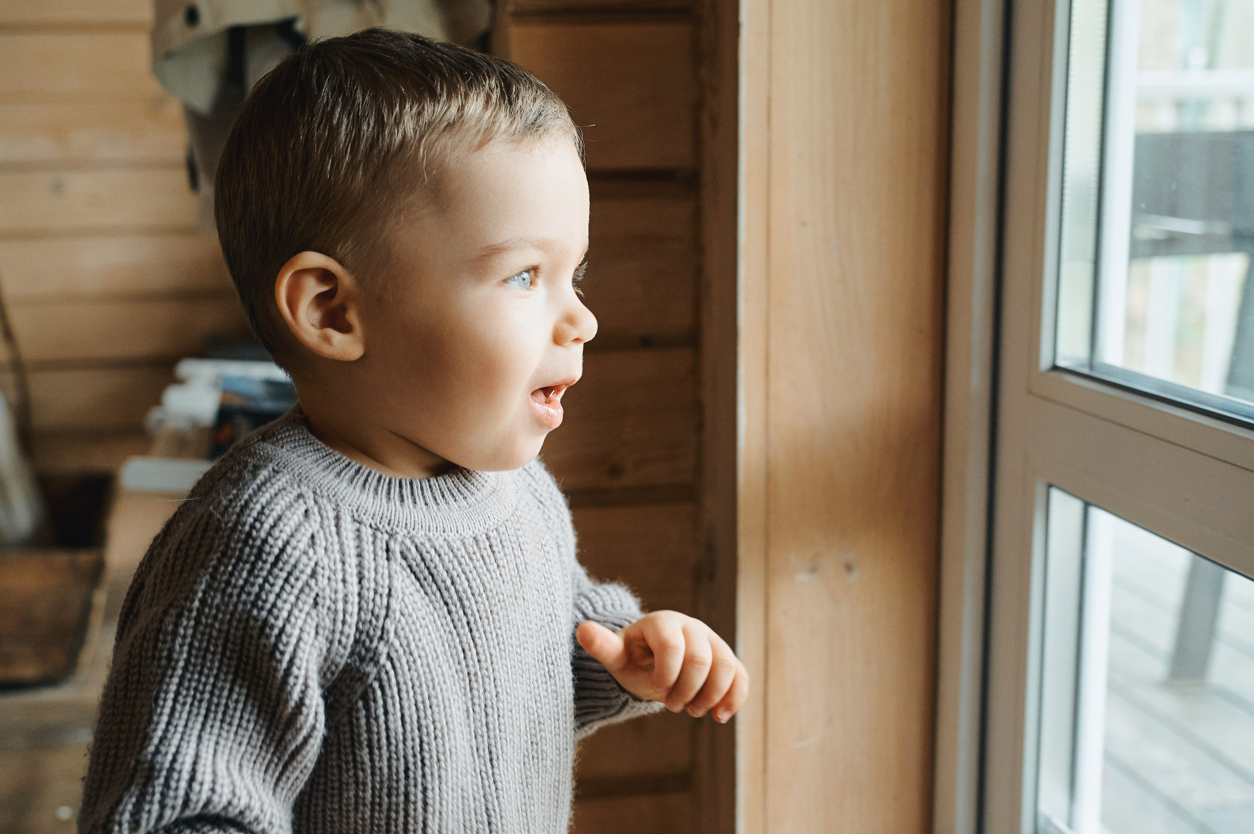 Ребёнок в вязаном свитере смотрит в окно с радостью и удивлением.