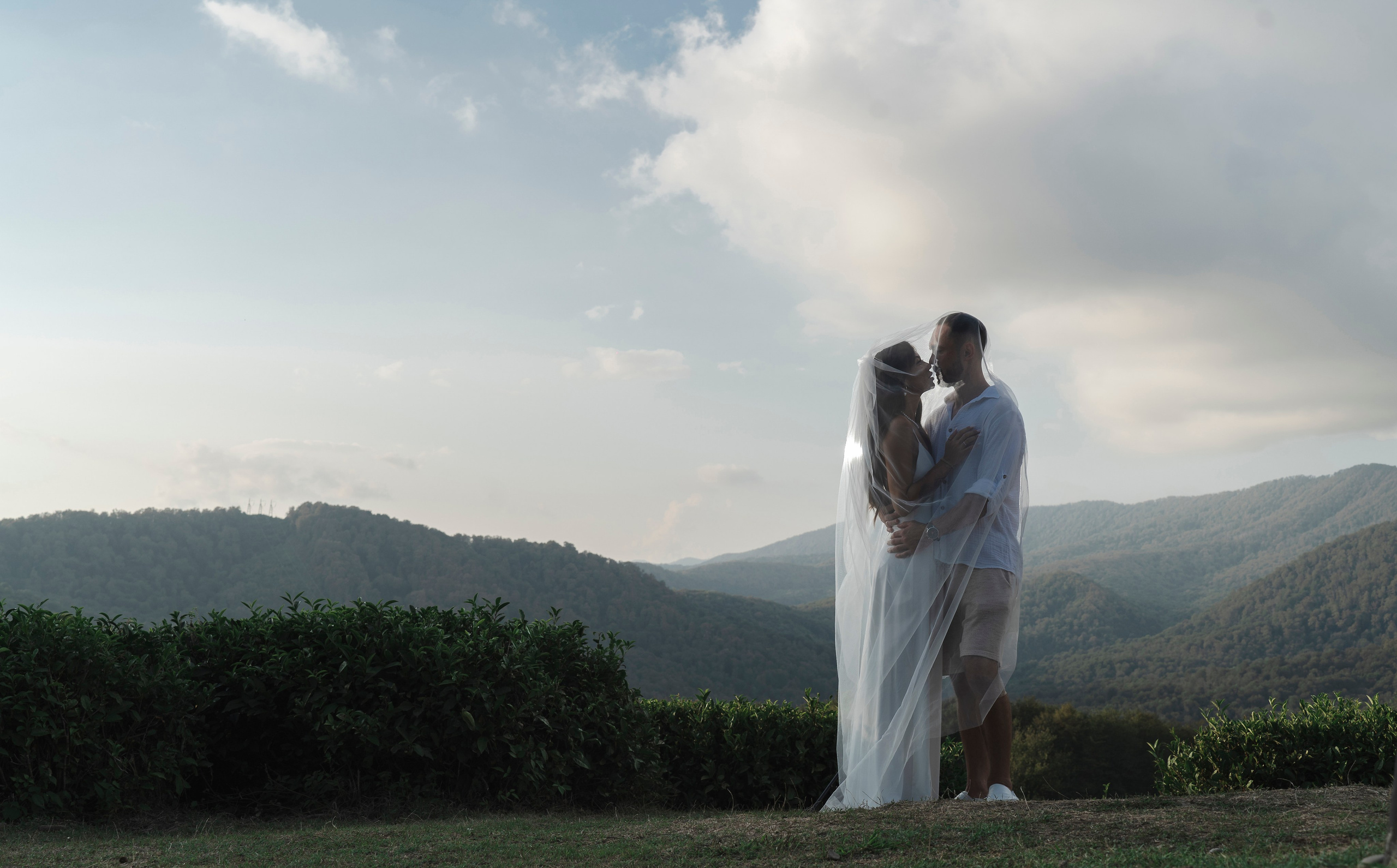 Мужчина и женщина, которые предположительно являются парой, стоят на чайном поле. На женщине белое платье и вуаль, а на мужчине белая рубашка и шорты. Женщина стоит лицом к мужчине, а мужчина - лицом к камере. Пара окружена пышными зелеными чайными растениями, а на заднем плане видны горы. Небо голубое с туманными облаками, и ярко светит солнце. Пара выглядит счастливой и влюбленной.