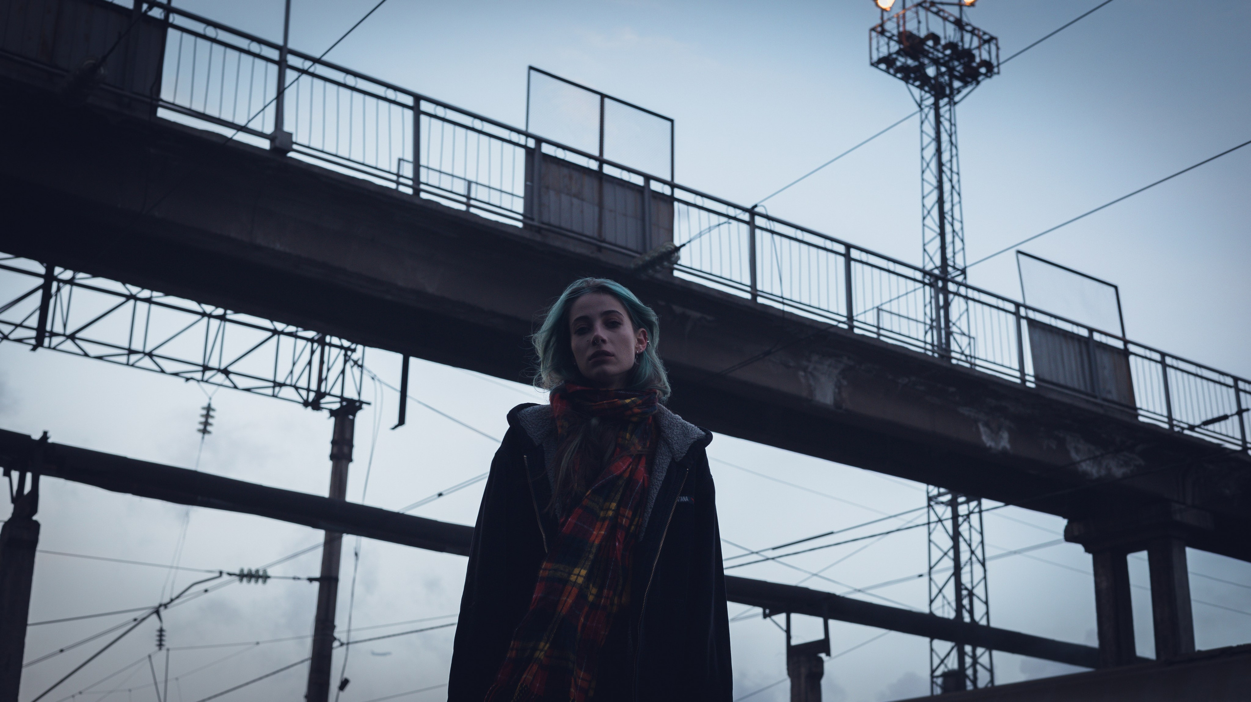 Девушка стоит под мостом, на фоне — городское небо и провода