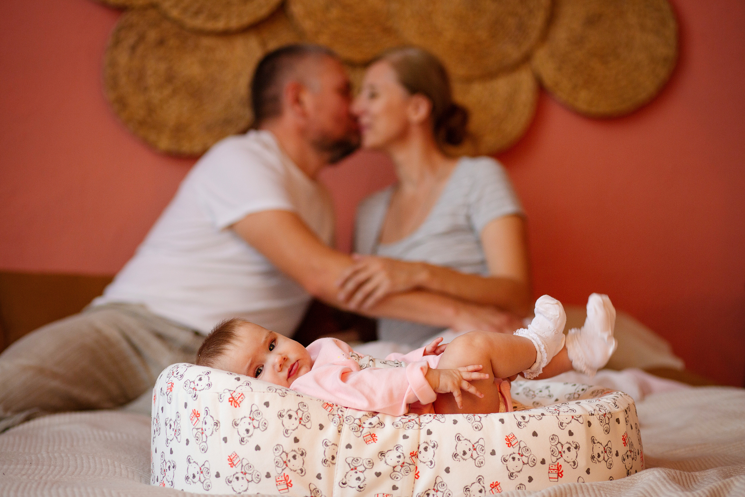 на переднем  плане   в переноске  лежит ребенок.  на  фоне  его родители  целуются.