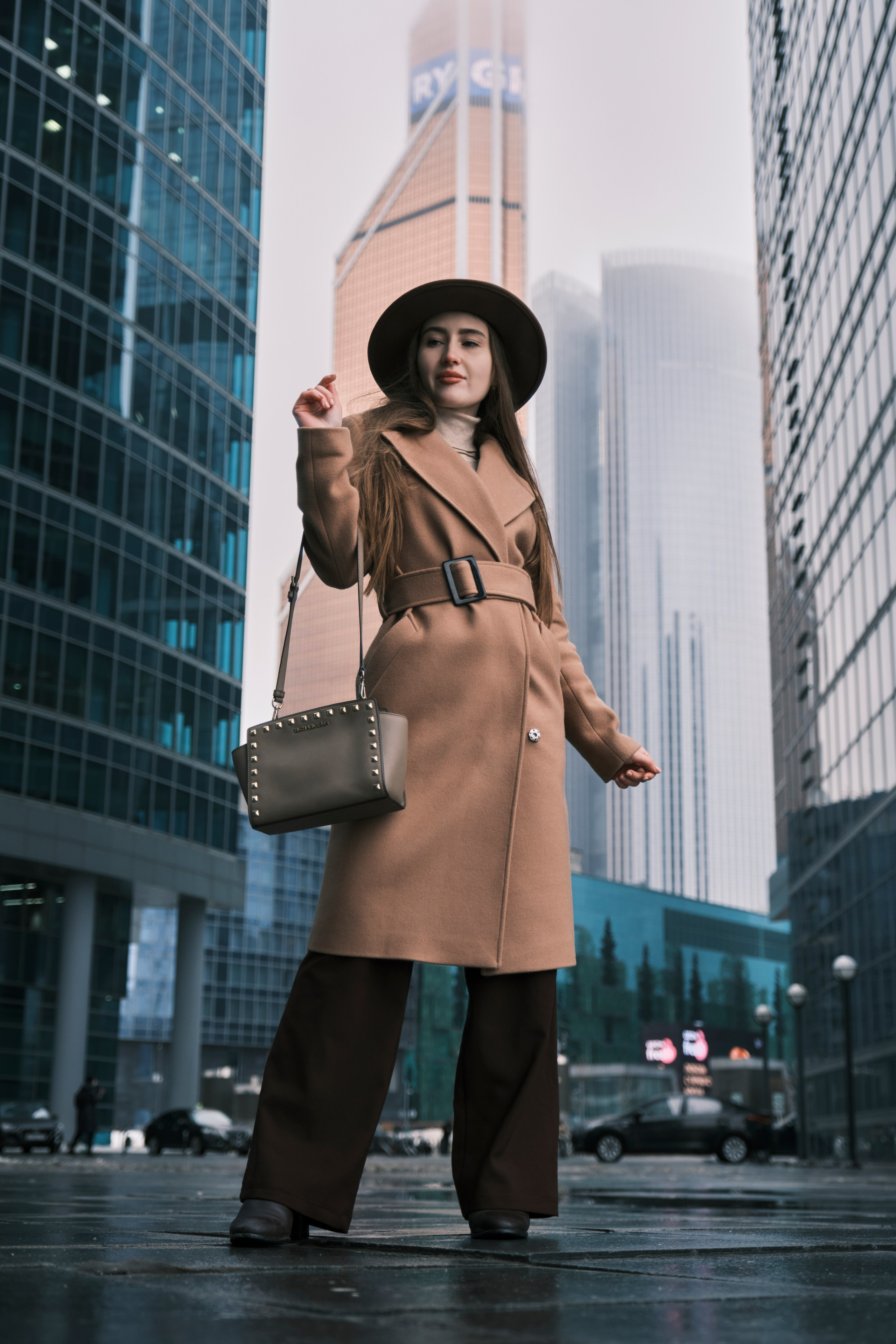 Женская фотосессия в Москва-Сити: Девушка в осеннем образе с маленькой сумочкой уверенно идет по улице на фоне высоких стеклянных зданий.