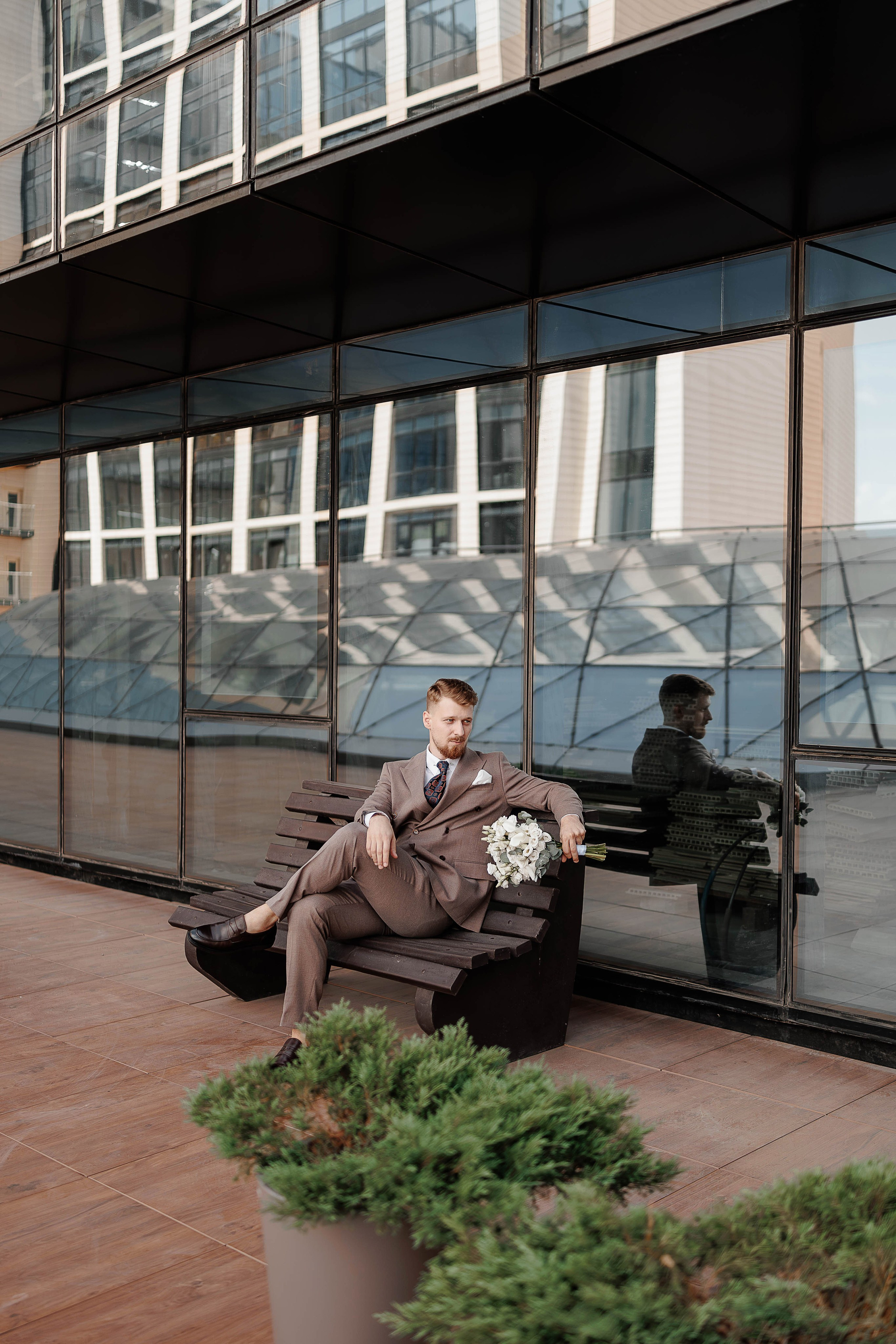 Жених сидит на лавочке с букетом перед фотосессией