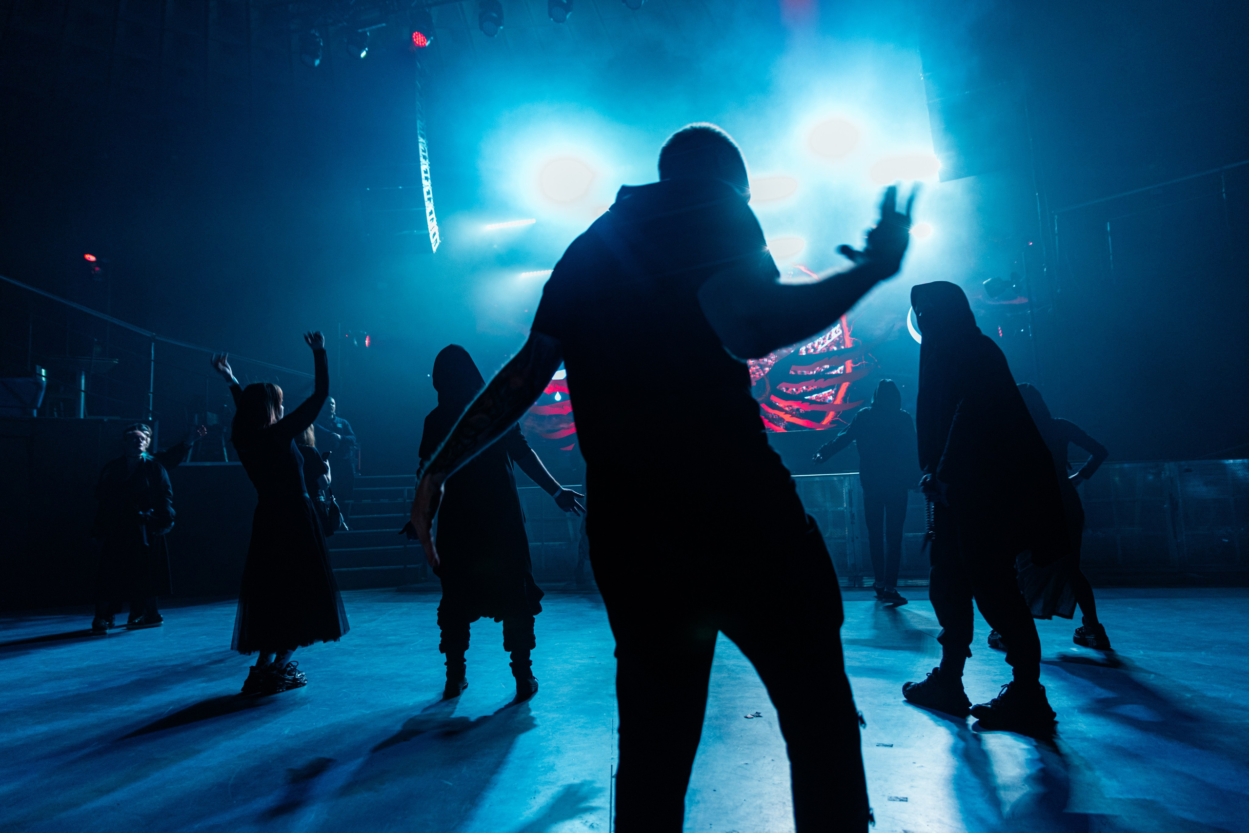 Фото из зала, силуэты людей со спины, на заднем плане ярко освещённая сцена, синий свет, от людей ложатся длинные тени, на переднем плане танцует человек