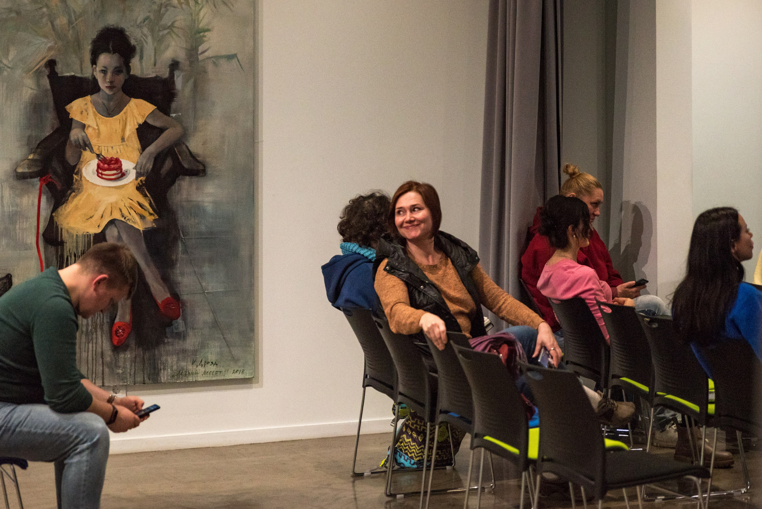 Лекционный зал, женщина сидит в последнем ряду, оборачивается и улыбается. На фоне большая картина, на ней девочка в кресле с тарелкой на коленях. В тарелке торт.