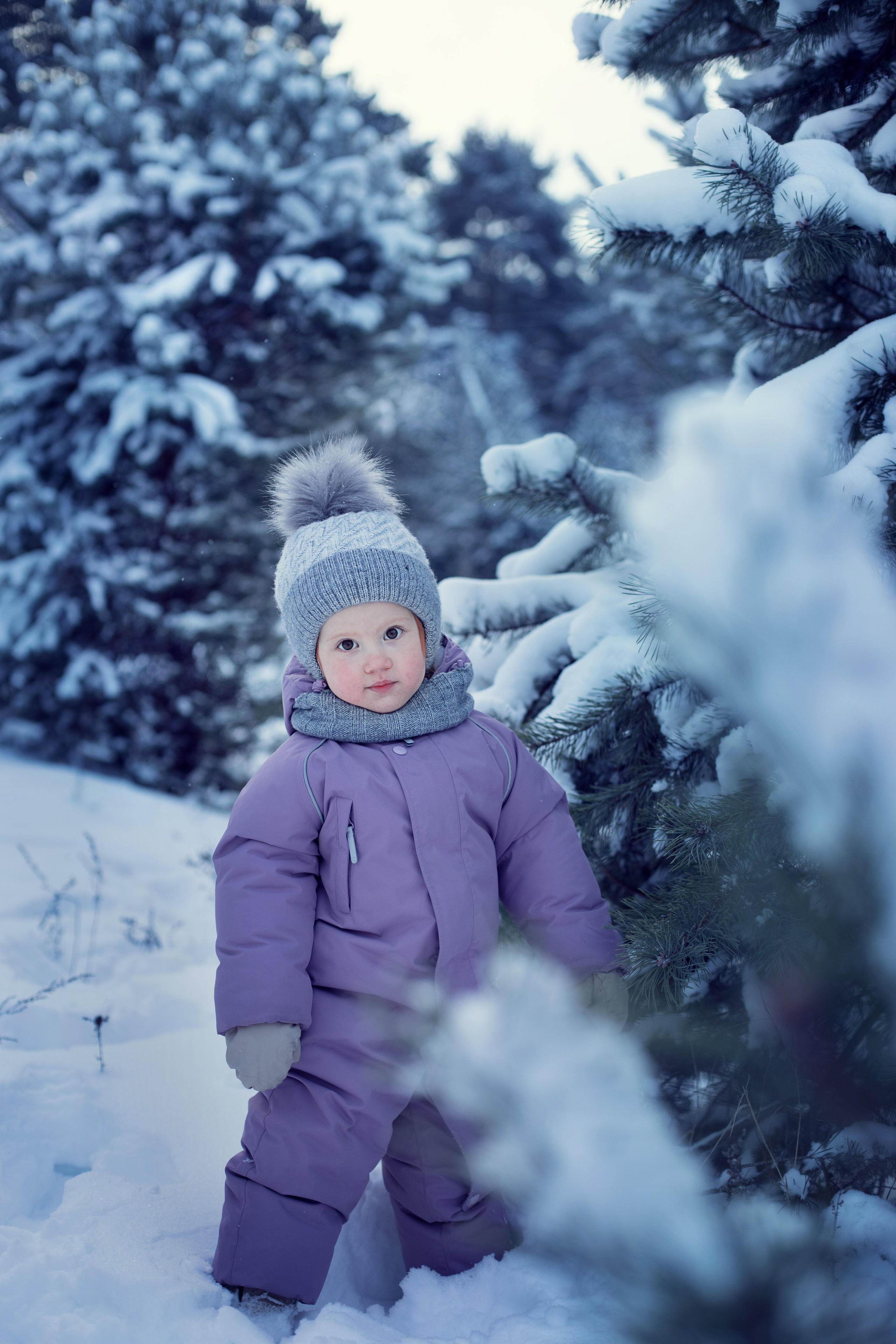 Девочка стоит в зимнем сосновом лесу в лиловом комбинезоне и серой шапке