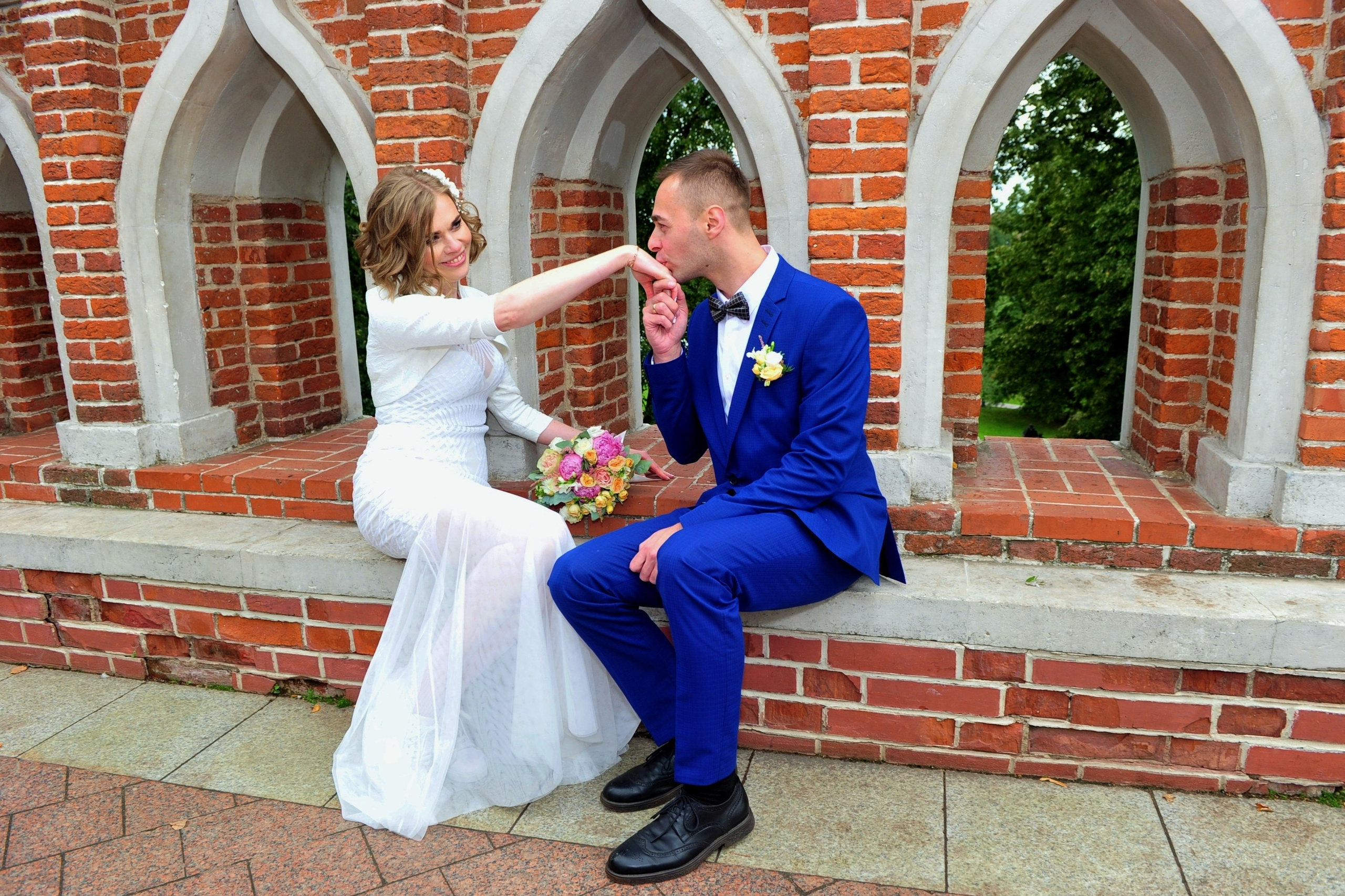 Свадьба в Царицыно хмурым прохладным летним днём, новобрачные сидят на мосту из красного кирпича в парке, жених целует ручку невесте