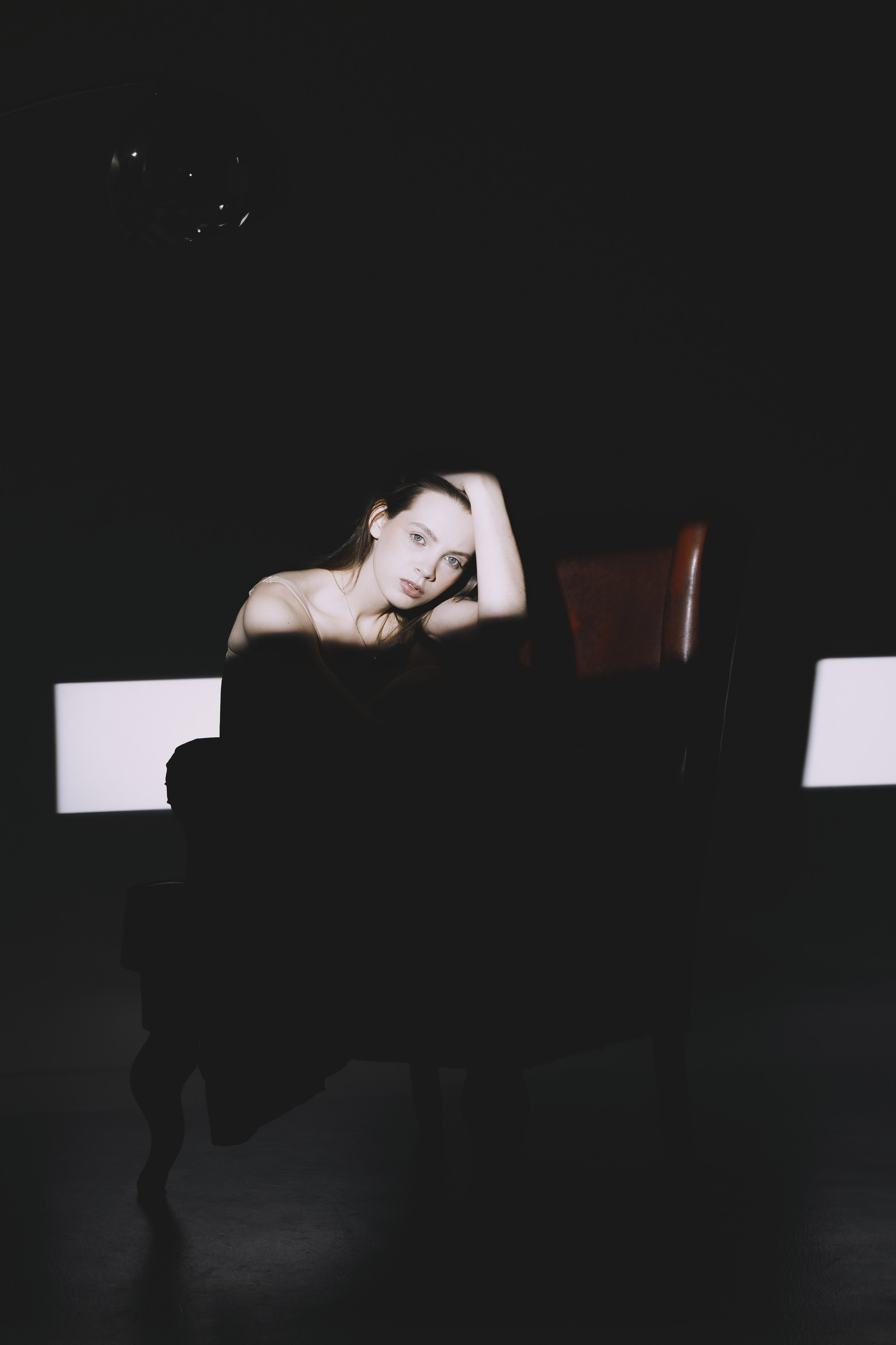 Атмосфера задумчивости в кадре: девушка в расслабленной позе на тёмном кресле. Игра света и тени создаёт глубокий, эмоциональный портрет.