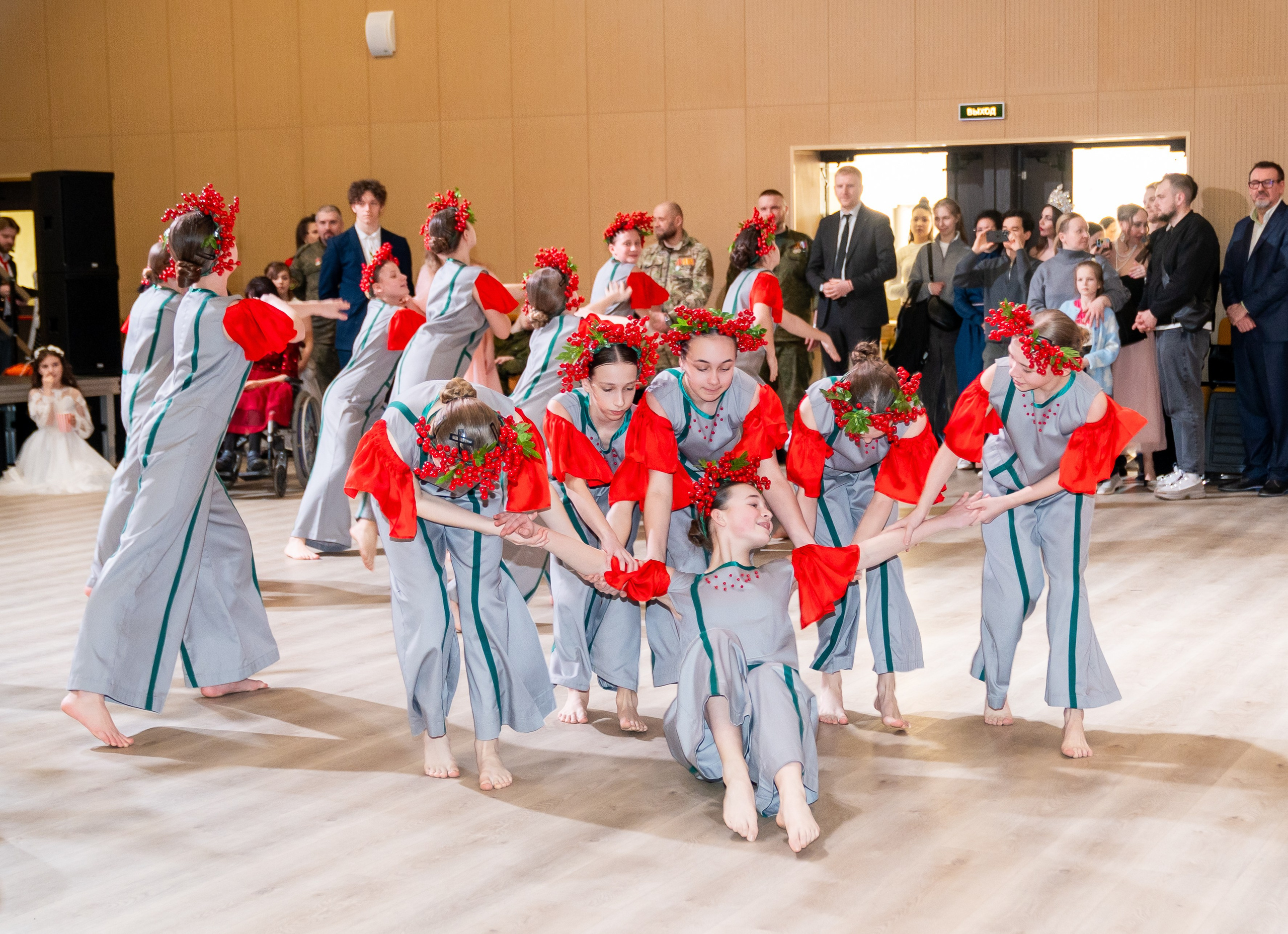 Танцевальная группа в серо-красных костюмах с красными венками позирует на сцене. На экране надпись «Мы все равны». Инклюзивный бал. Фотограф Мальцев Евгений.