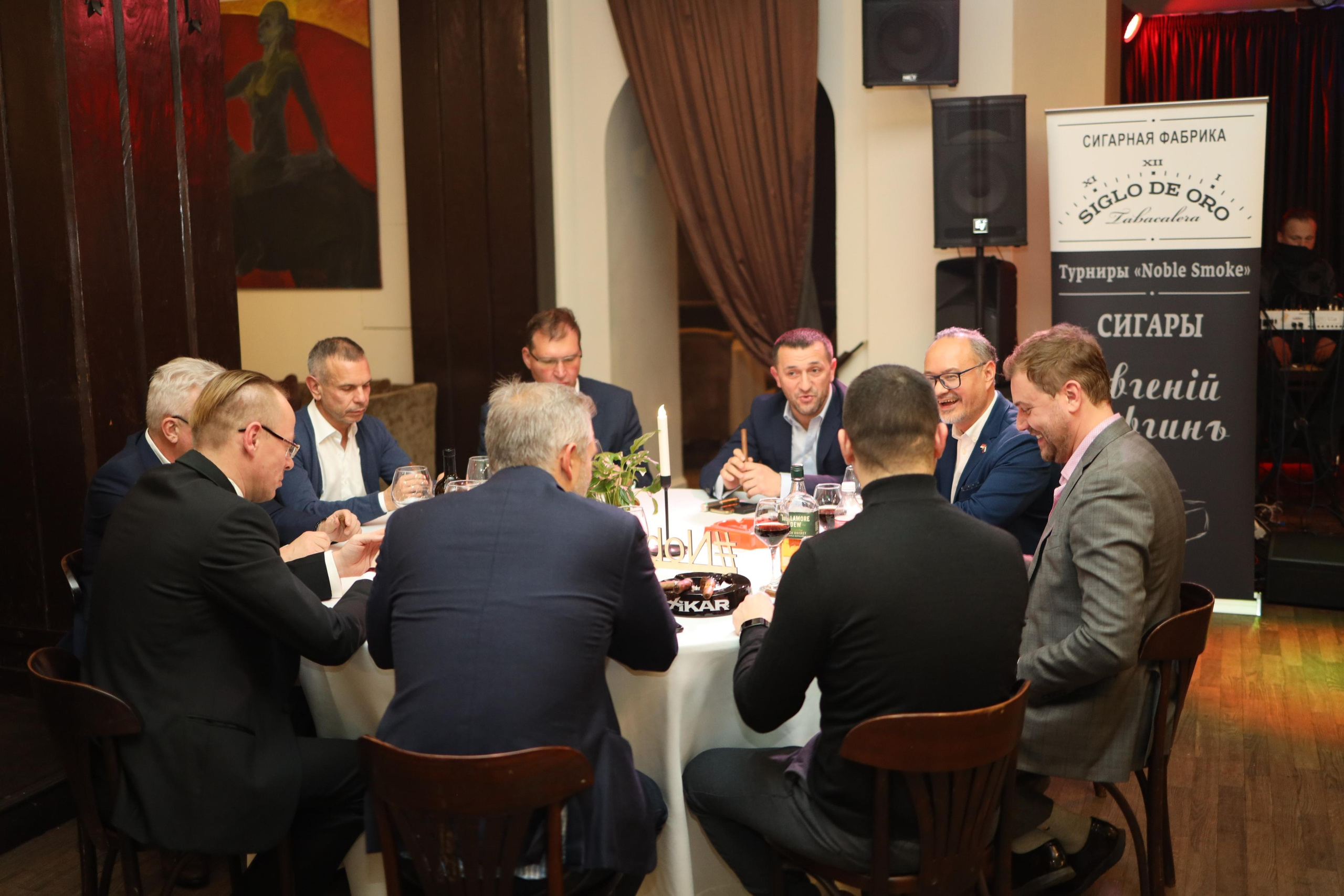 Мужчины сидят на круглым большим столом и курят сигары.