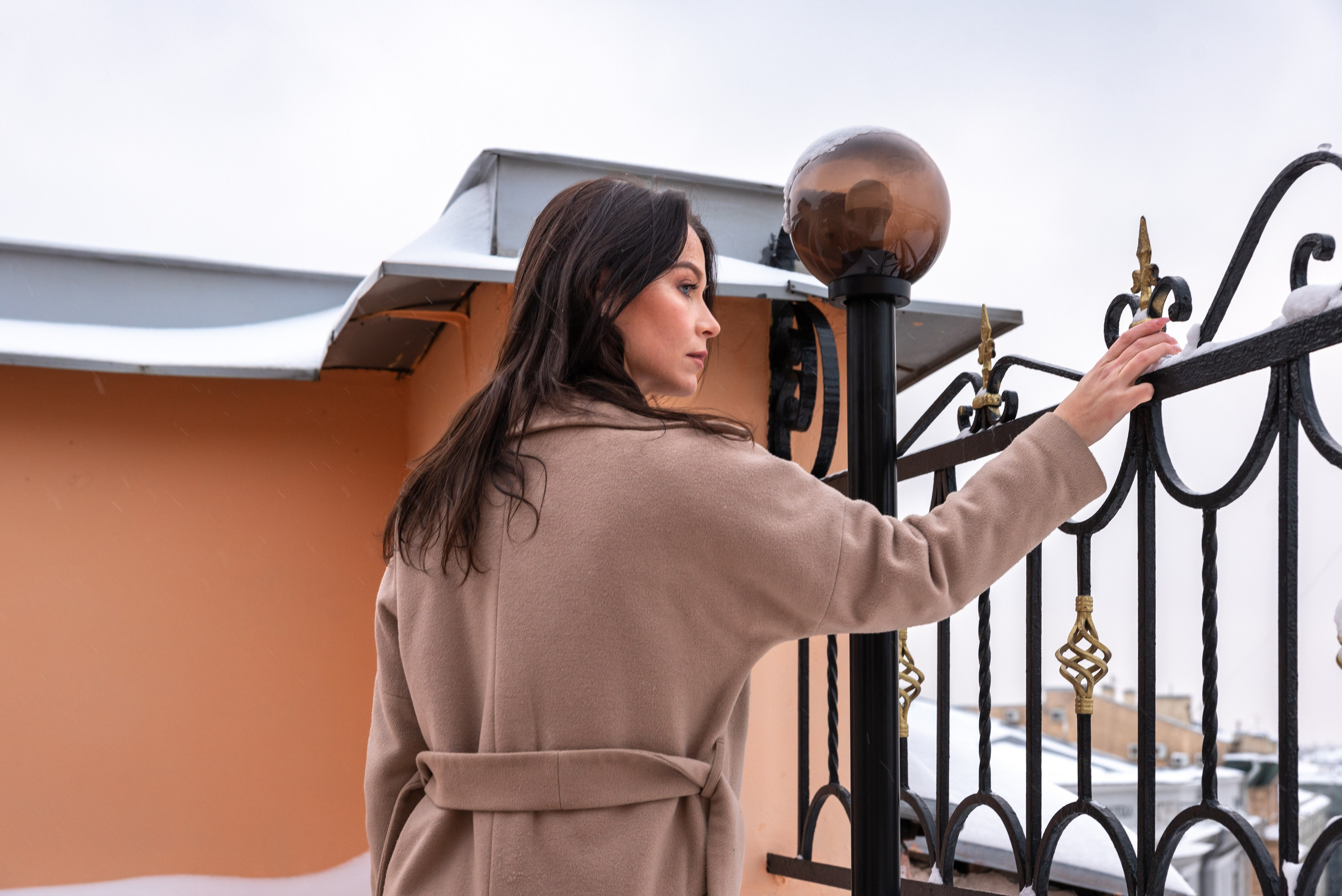 Девушка с длинными волосами в бежевом пальто стоит у металлического ограждения на крыше здания. Дотрагивается рукой до решетки.