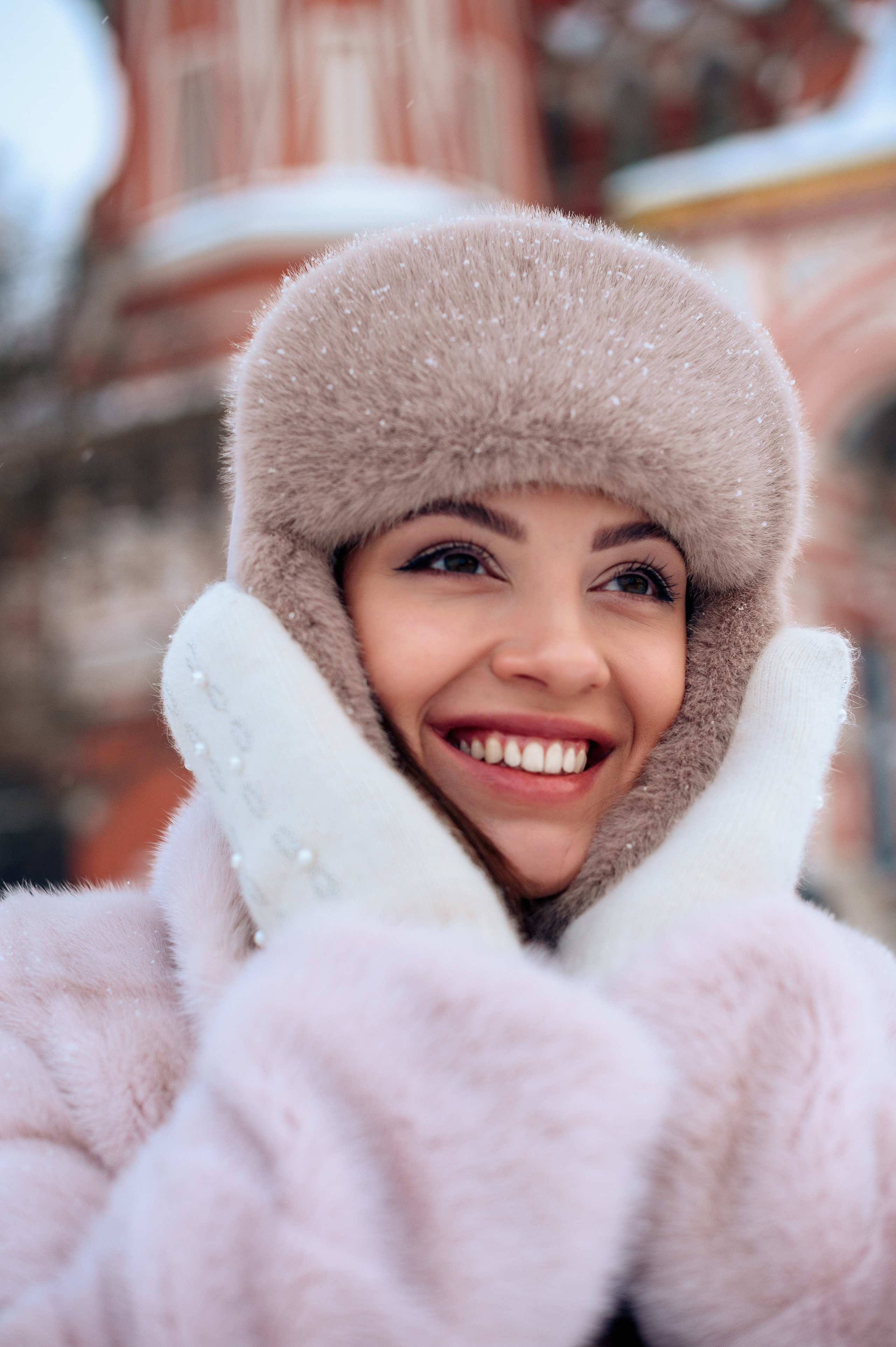 Портрет девушки крупным планом с красивой белоснежной улыбкой, которая прижимает свою шапку к щекам. На ее руках белые варежки