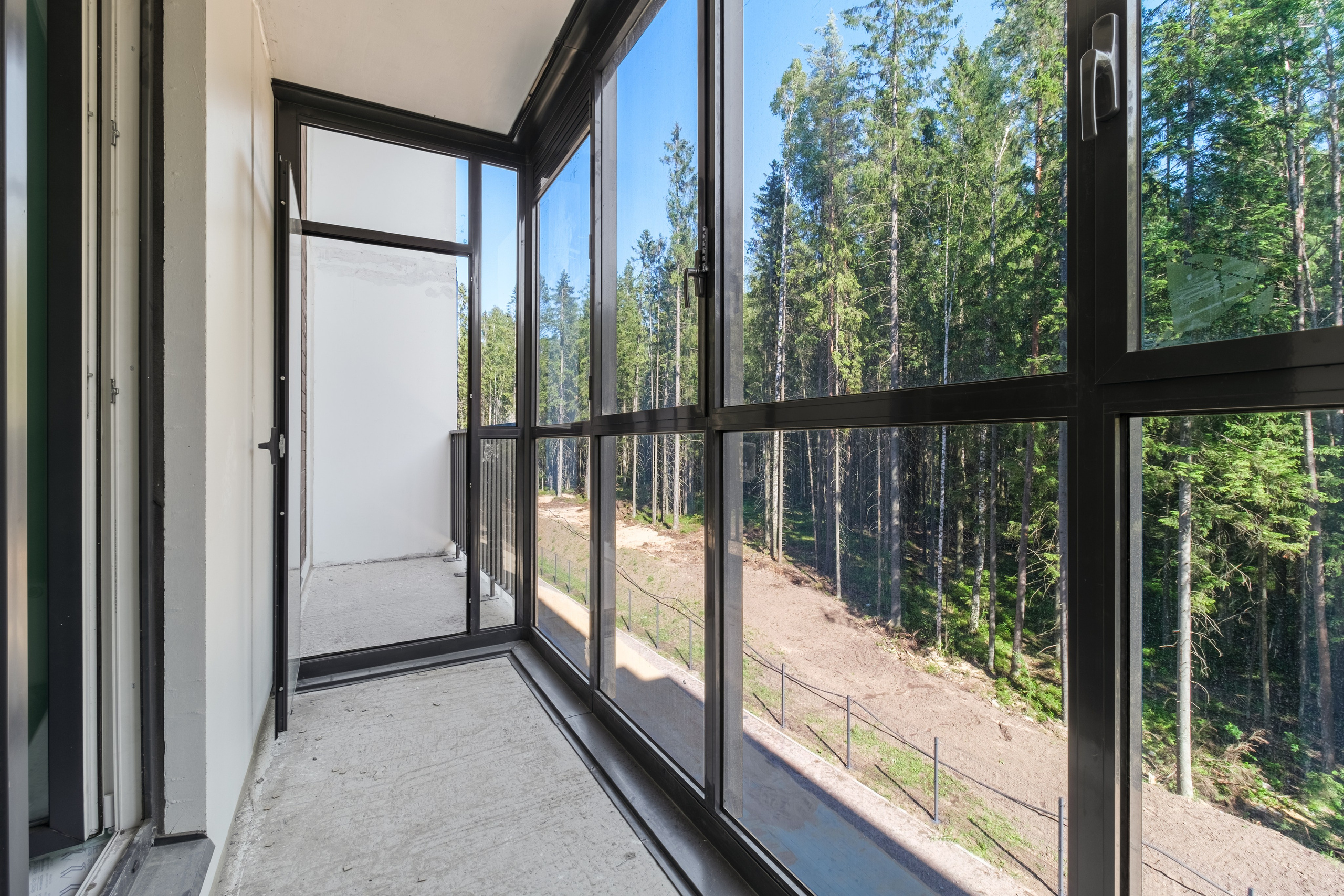 Балкон с большими окнами от пола до потолка, через которые виден лес. Окна оснащены металлическими рамами.