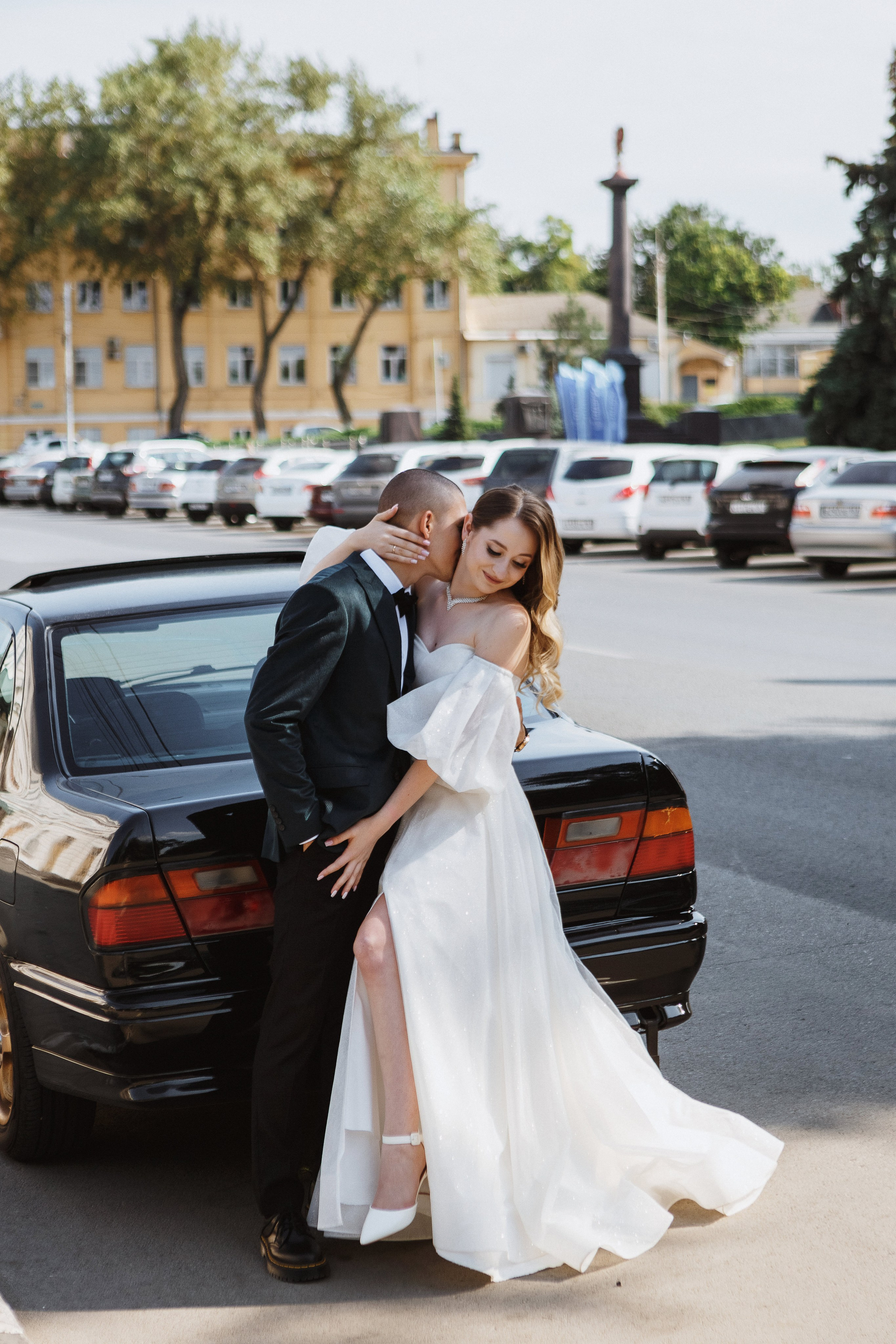Свадебная фотосессия в Таганроге: жених в тёмном костюме обнимает невесту в белом платье, которая сидит на багажнике автомобиля. На заднем плане улица Петровская с припаркованными машинами и памятником на площади.