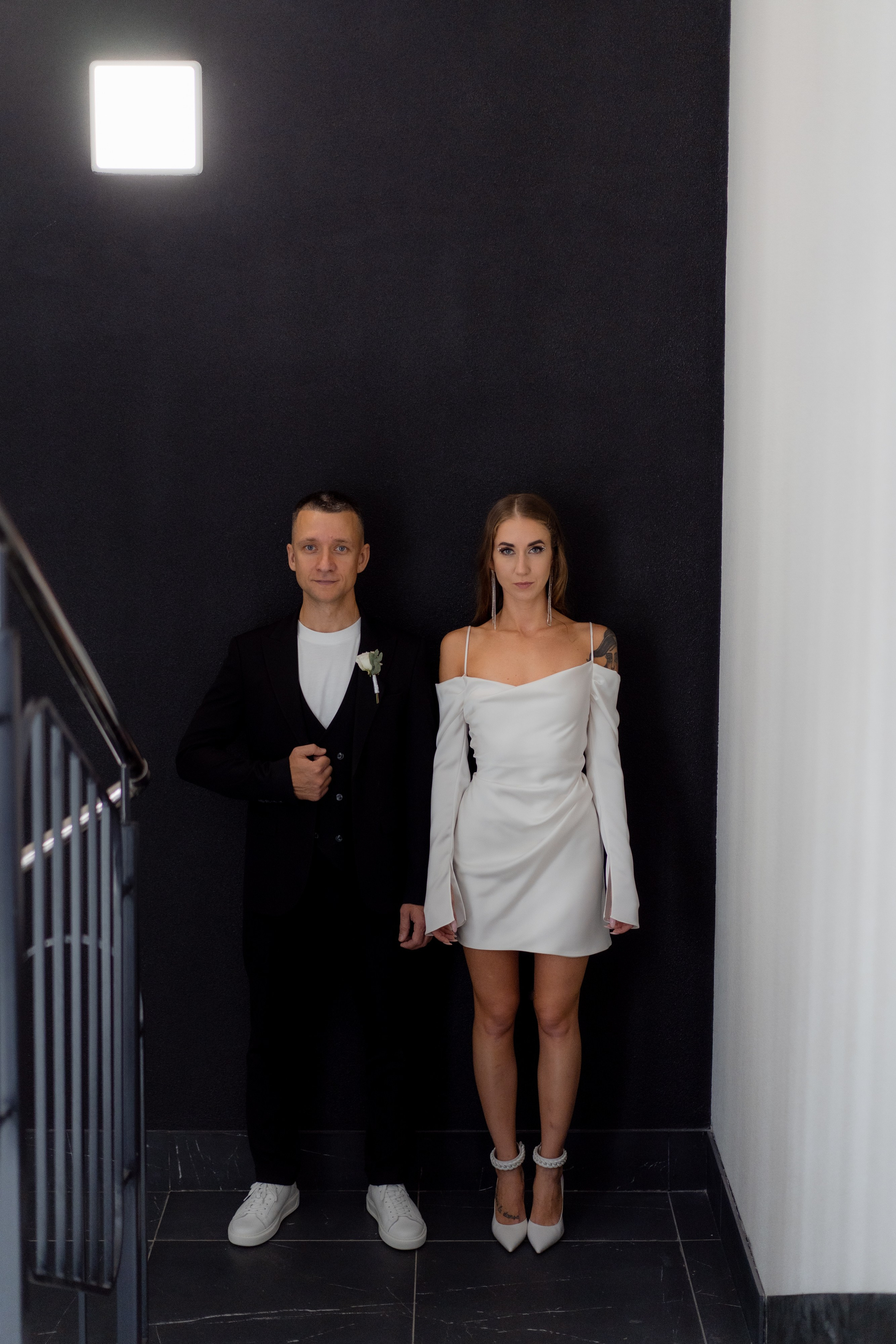 Невеста с татуировками и в коротком белом платье и жених в черном костюме стоят на черной лестнице