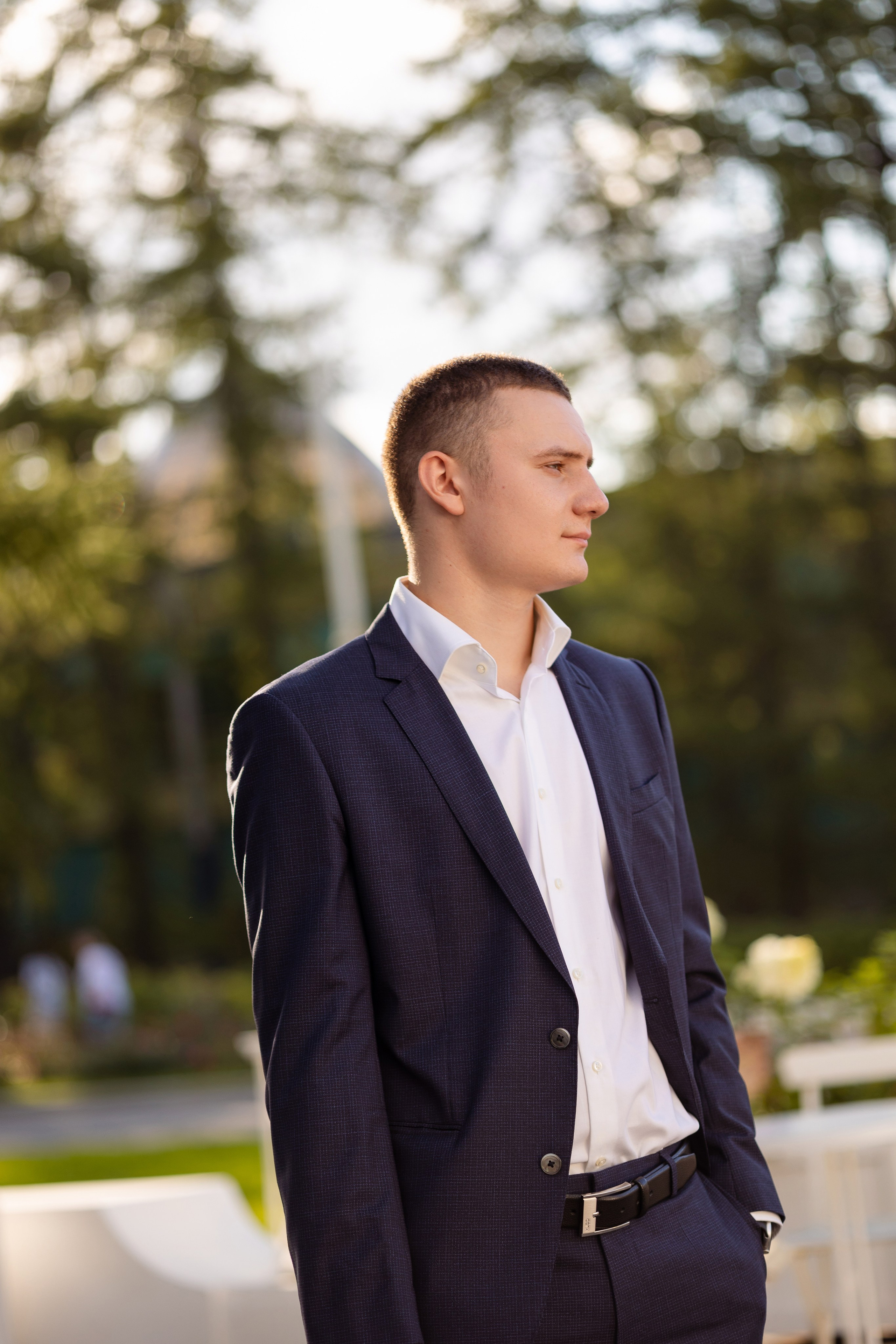 На улице, на фоне деревьев жених стоит в пол оборота.