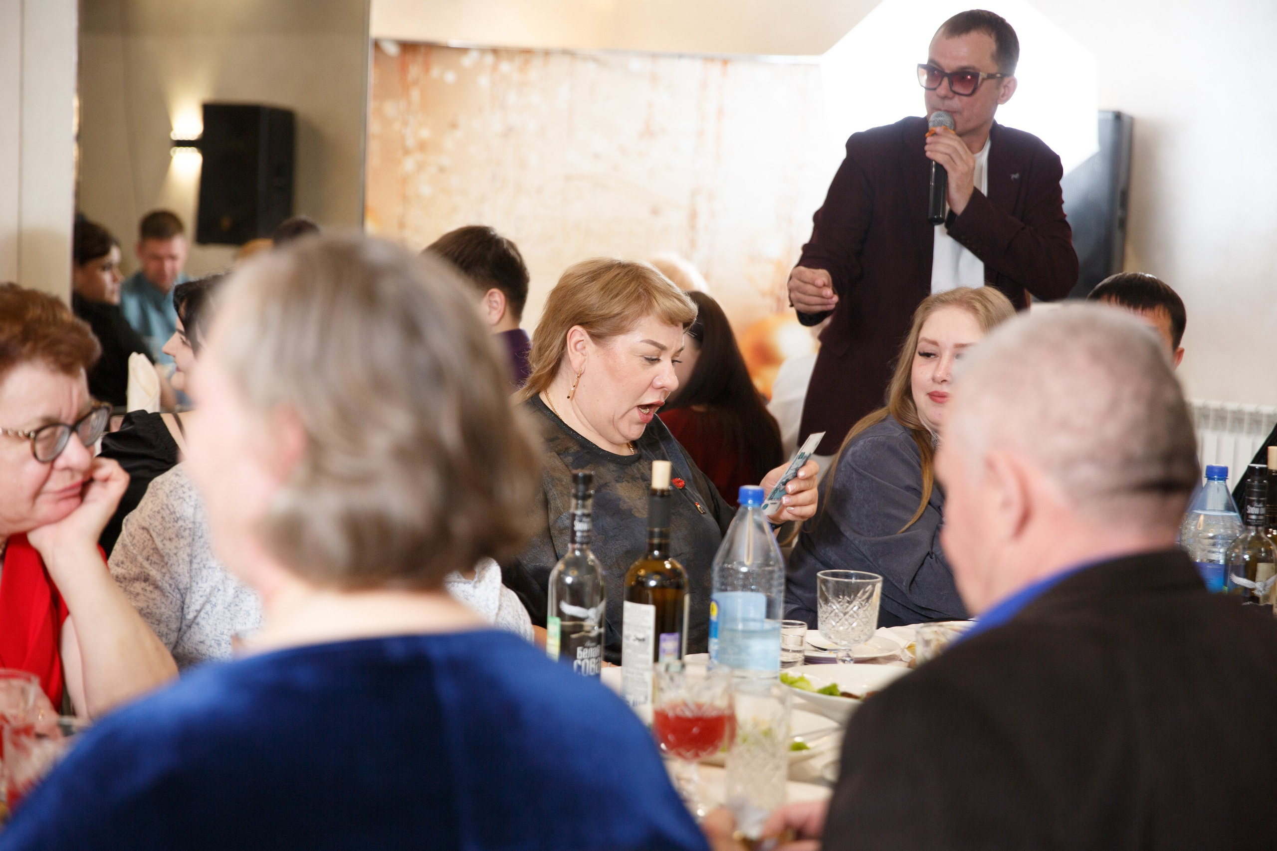 Мужчина с микрофоном, гости аплодируют и улыбаются.