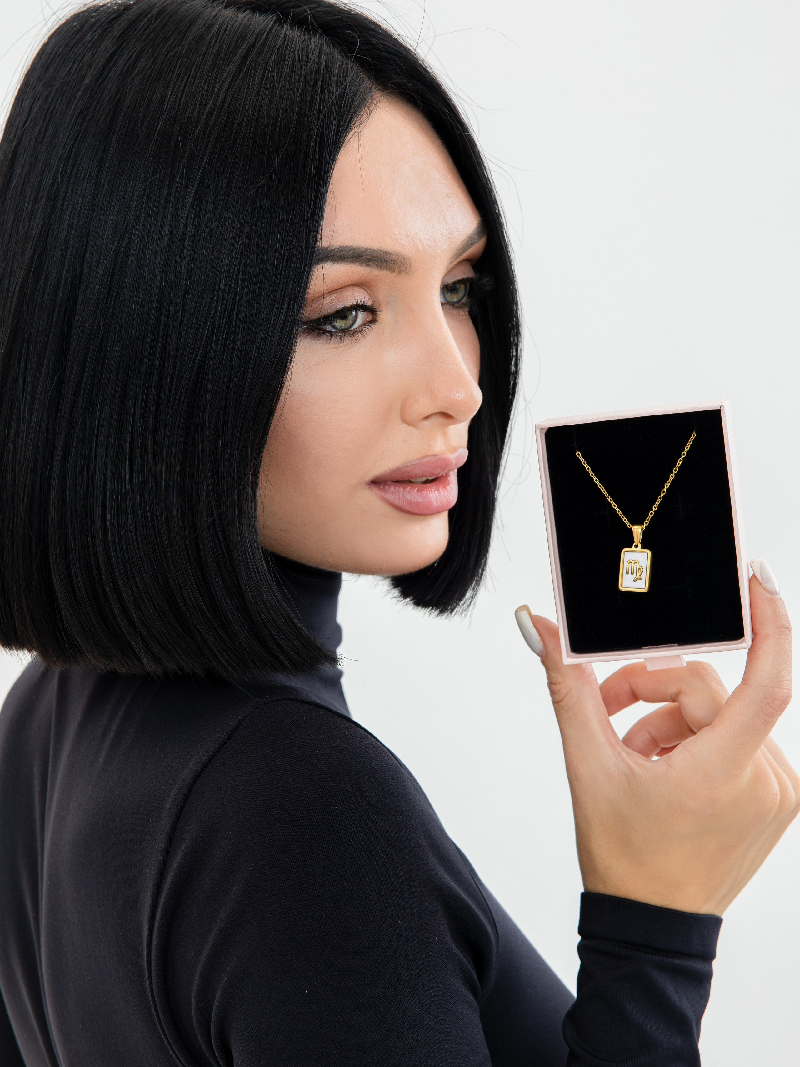 Портрет девушки брюнетки с зелеными глазами в черной водолазке держащую коробочку с золотым украшением в руке на белом фоне