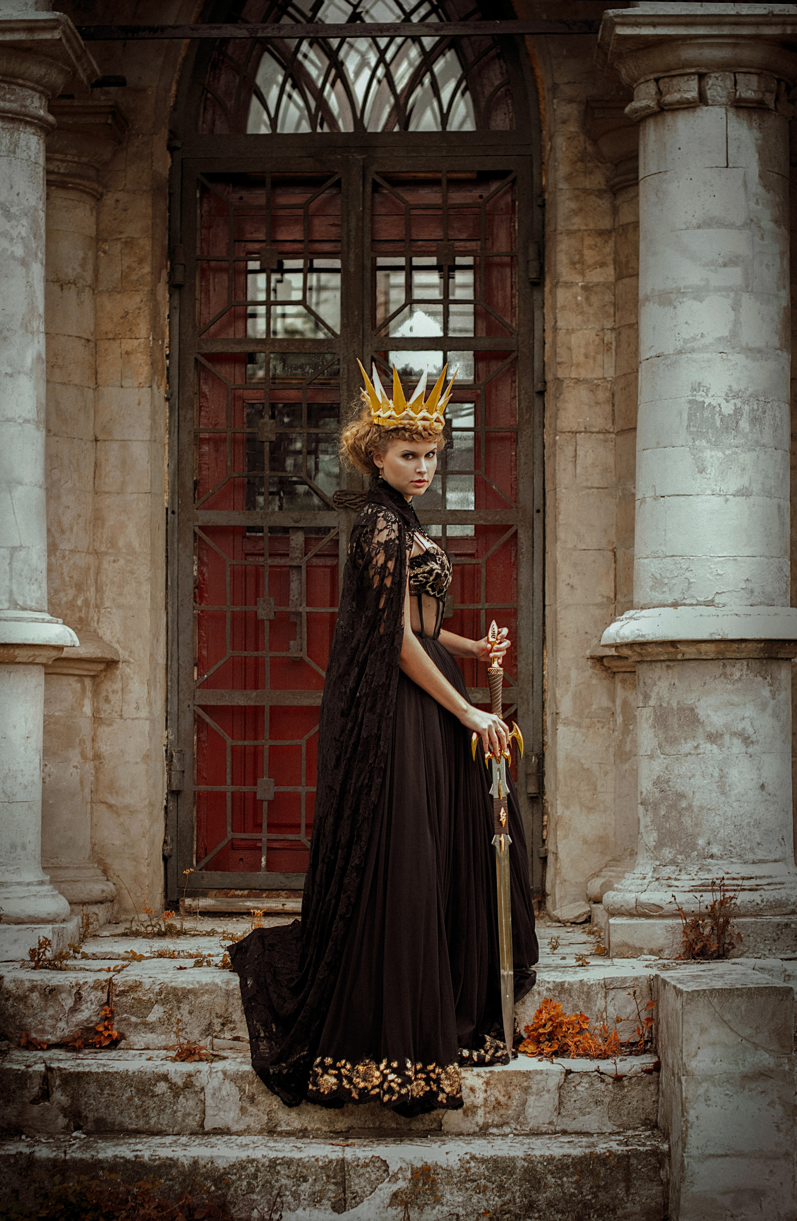 Женщина в царственном черном платье и золотой короне стоит на каменных ступенях, держа в руках меч. На заднем плане — богато украшенный, состаренный дверной проем, передающий средневековую и драматическую атмосферу