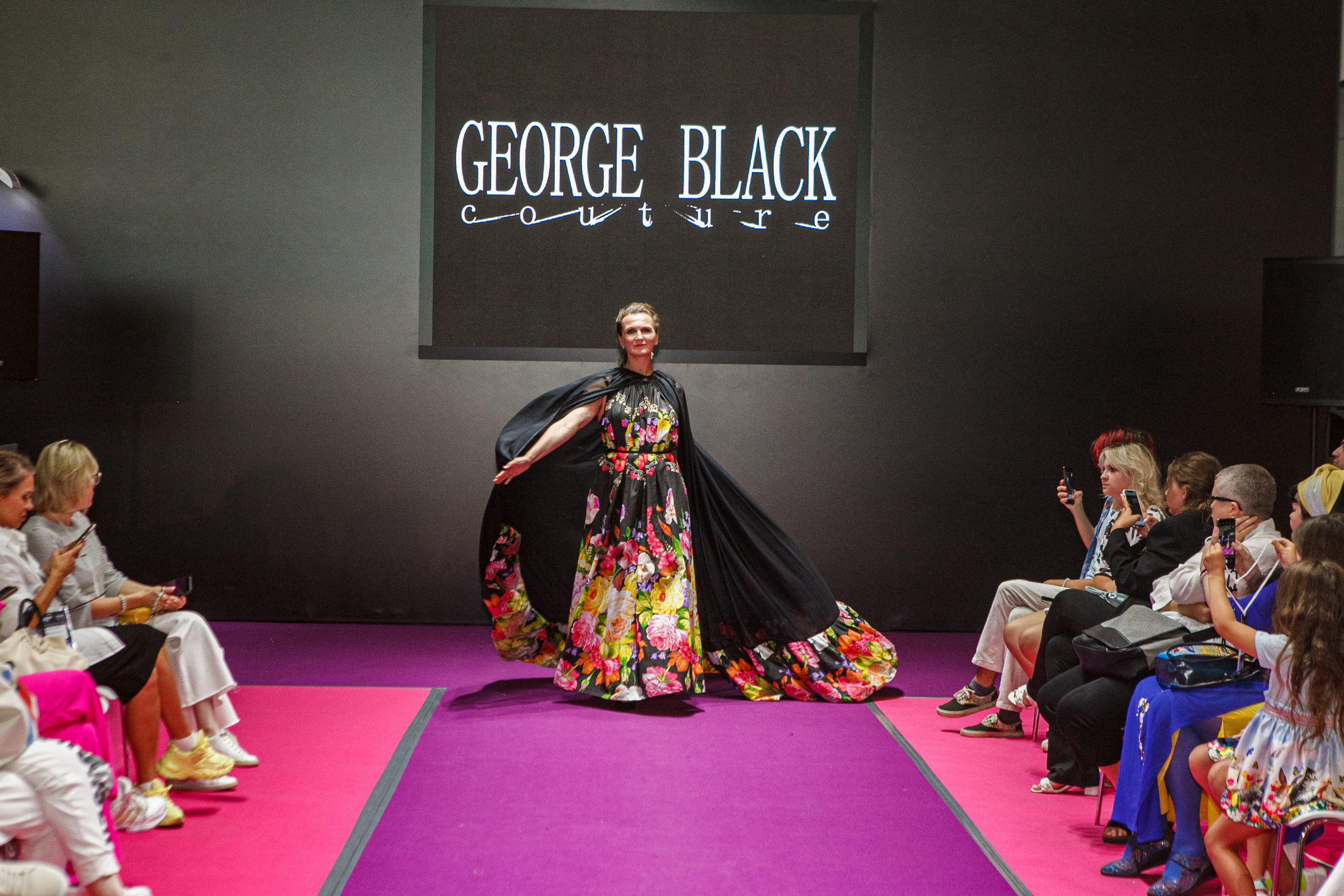  "Модели в ярких нарядах проходят по подиуму во время показа мод, освещенные яркими софитами и окруженные восторженными зрителями."