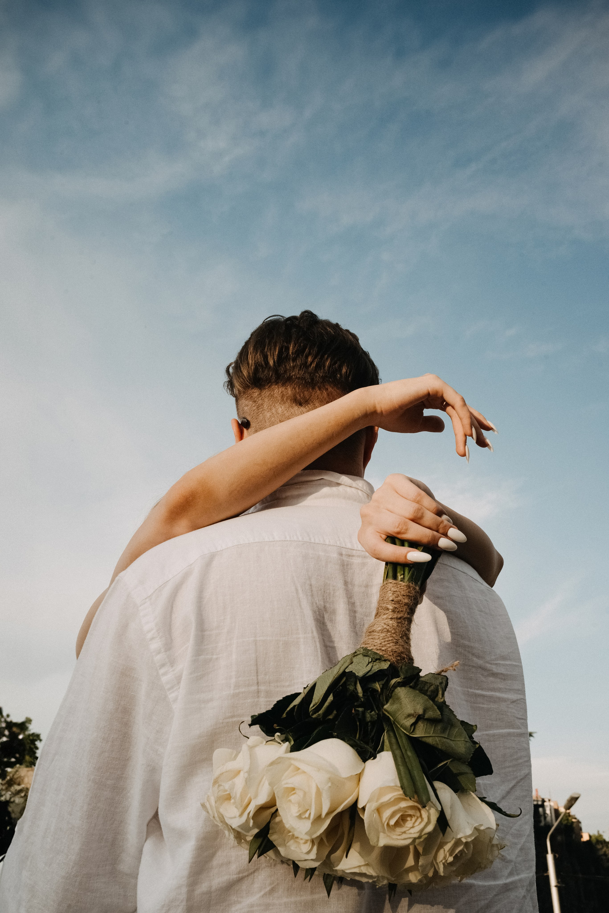 Женщина обнимает мужчину, держа в руках свадебный белый букет цветов. Мы видим только спину жениха