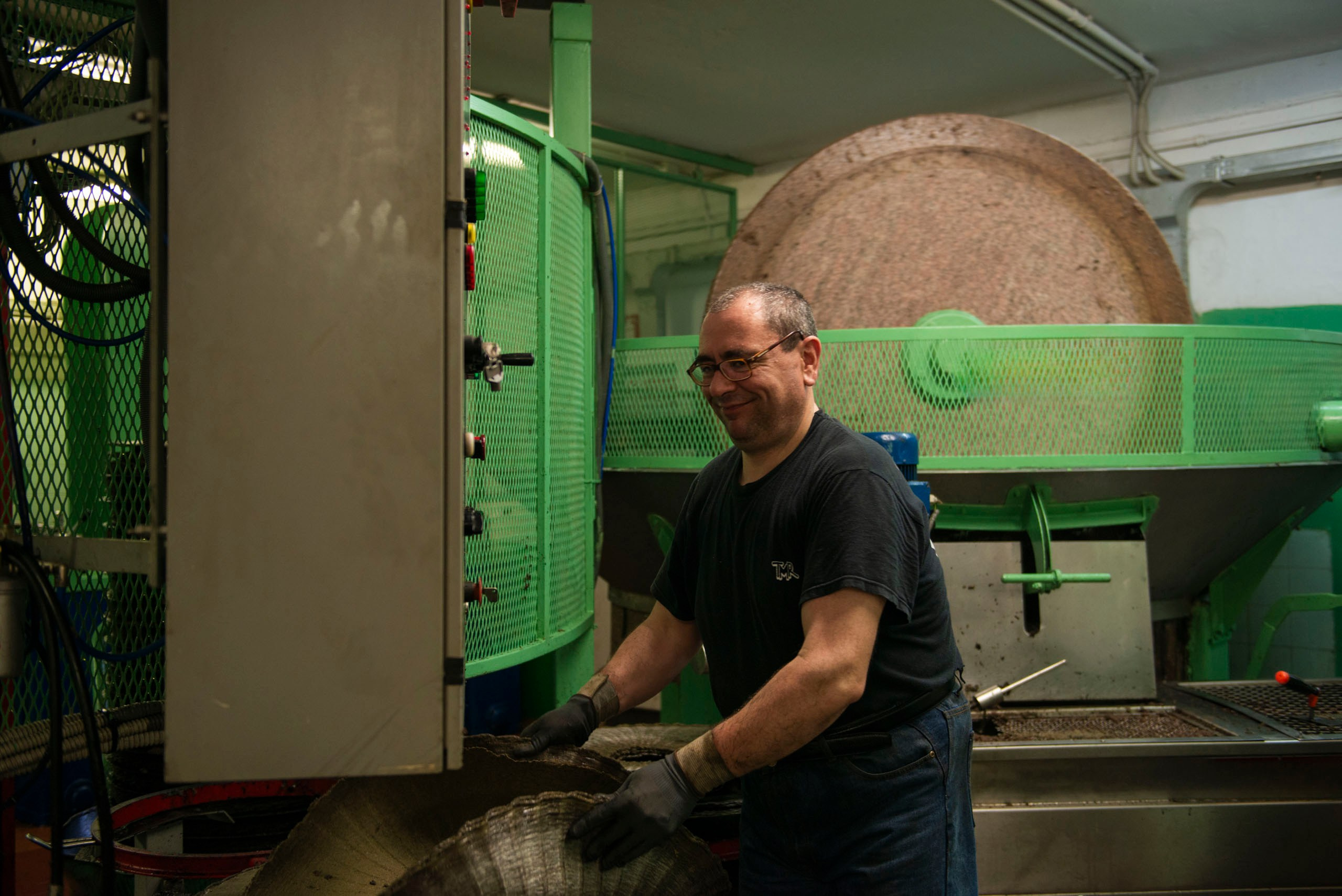  Улыбающийся мужчина в черной футболке и перчатках вручную укладывает плетеный фильтр (скуртину) в традиционный гидравлический пресс для производства оливкового масла.