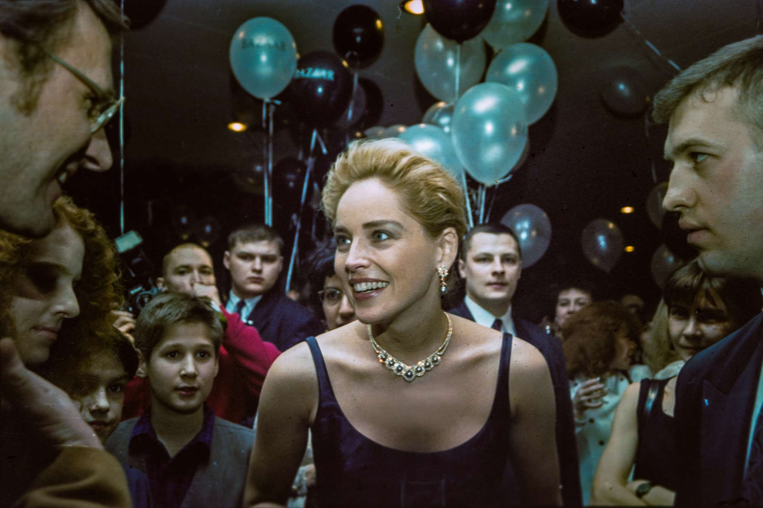 Женщина в вечернем платье среди толпы на светском мероприятии, воздушные шары, вспышки камер, репортажная фотография.                 Woman in an evening dress surrounded by a crowd at a social event, balloons, camera flashes, reportage photography.