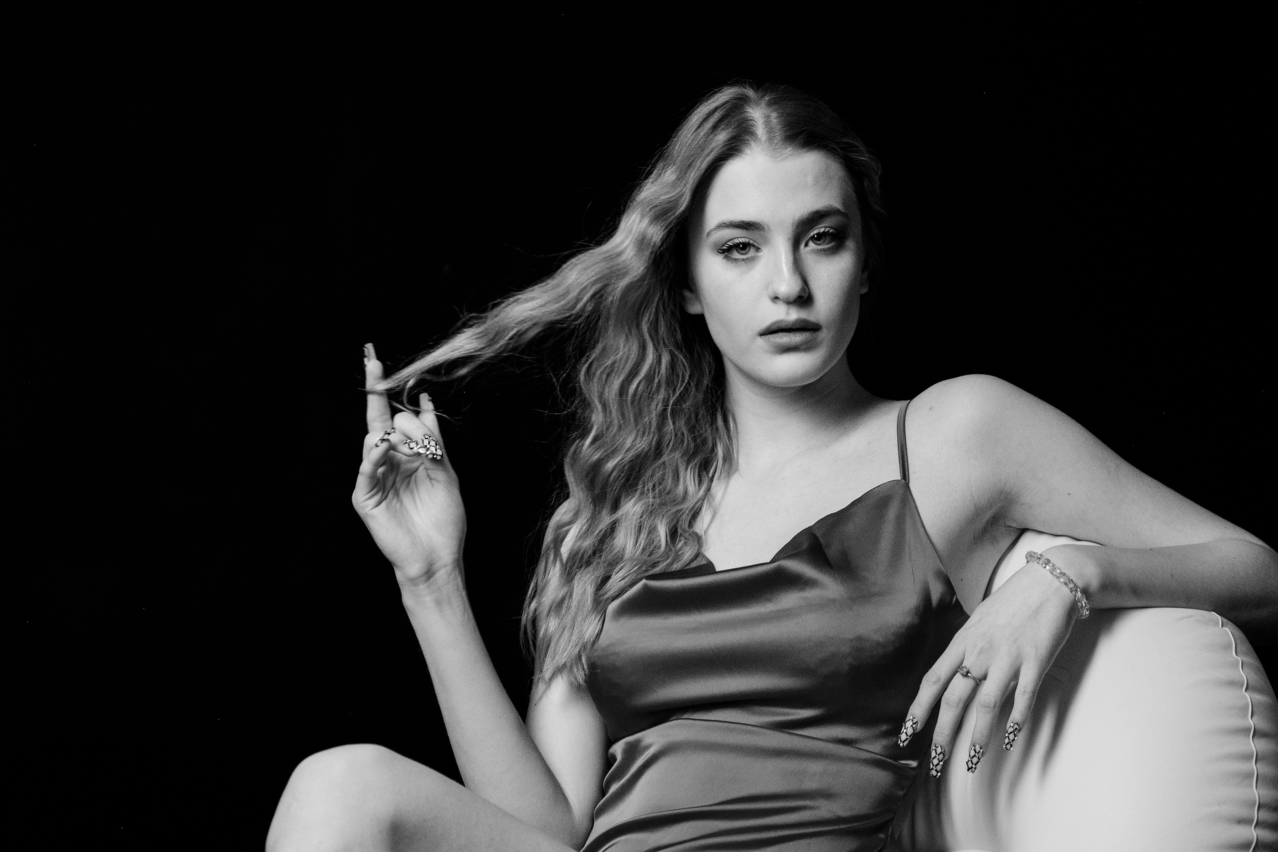 Светловолосая девушка сидит в кресле в свободной позе,положив левую руку на спинку,а правой рукой играет с прядью волос..