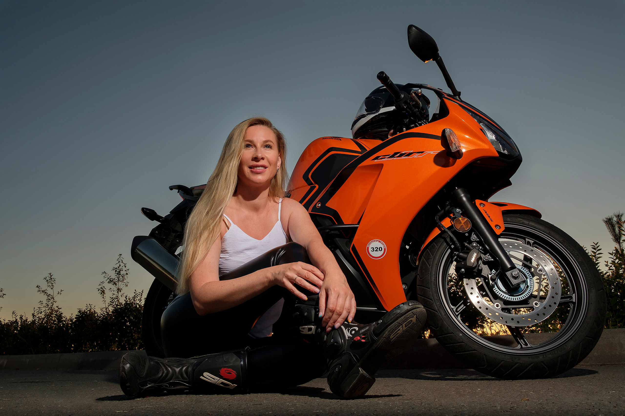 Блондинка сидит возле своего байка на фоне закатного неба.