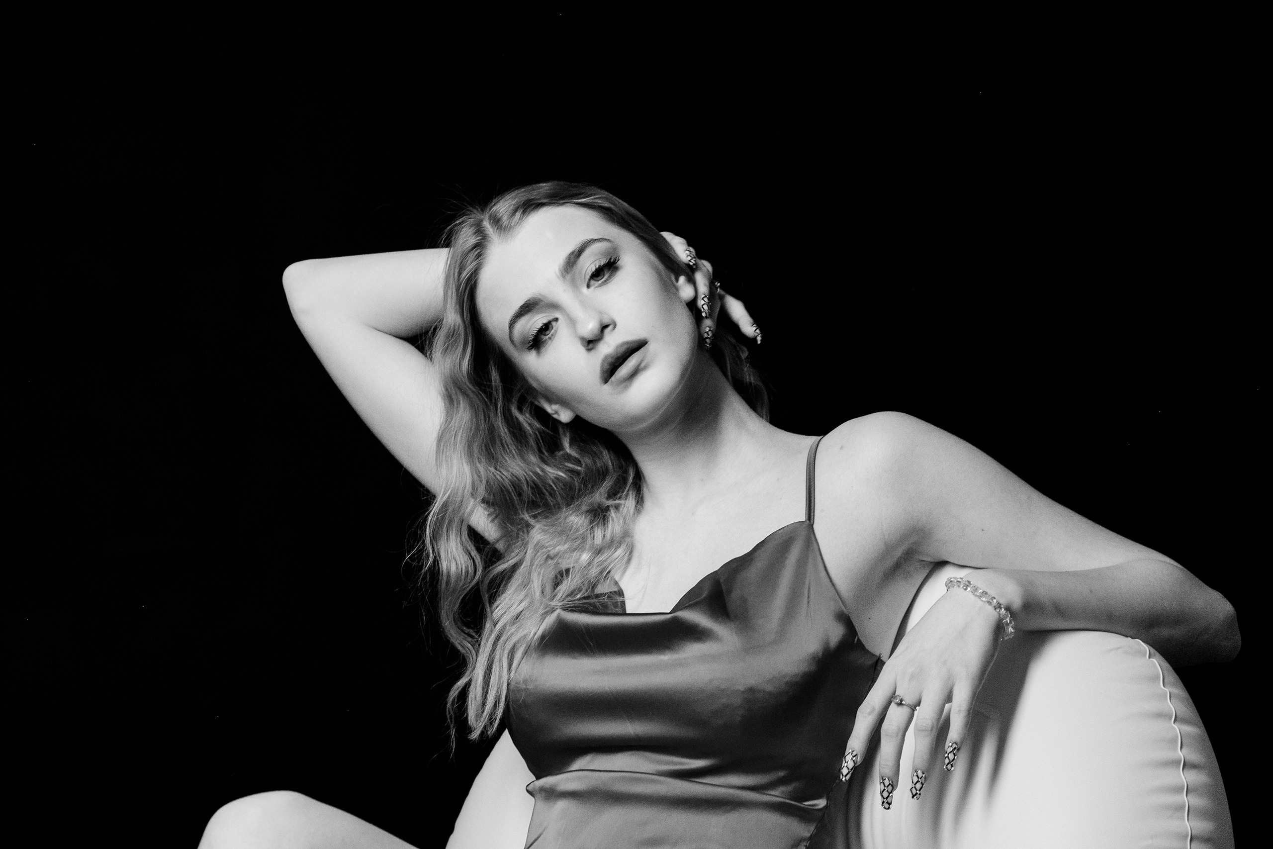 Светловолосая девушка сидит в кресле в свободной позе,правая рука закинута за слегка наклоненную голову,а левая лежит на спинке кресла..