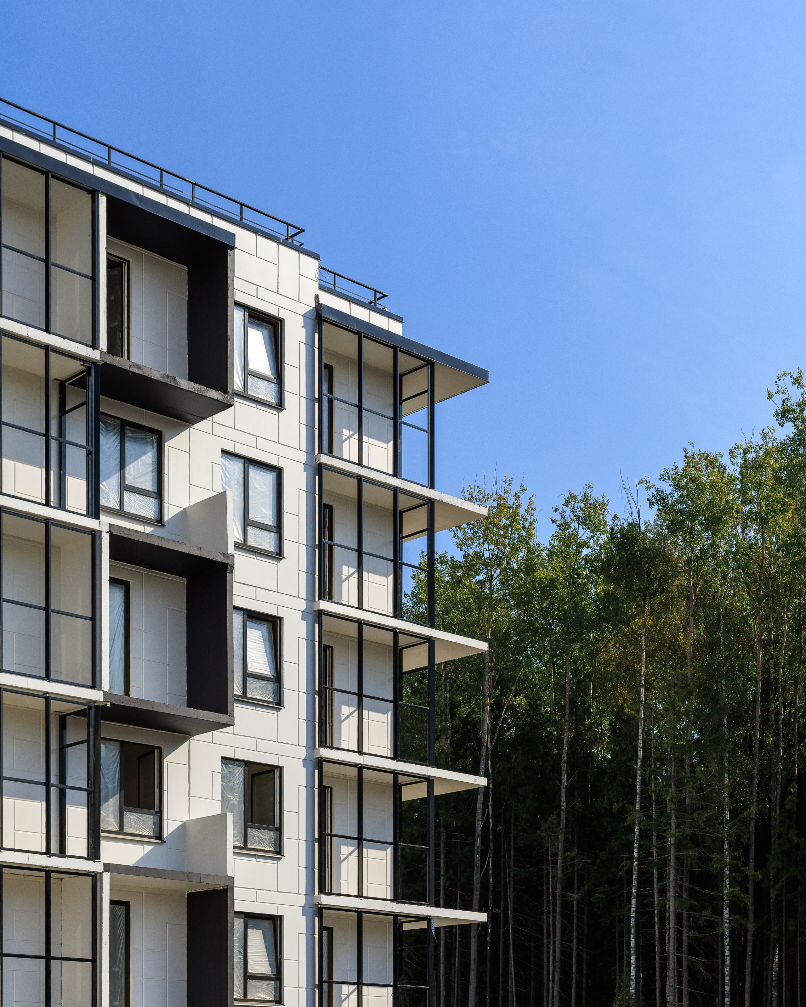 На изображении представлено современное многоэтажное здание, окружённое зелёной растительностью. Здание выполнено в современном архитектурном стиле с использованием стекла и бетона.