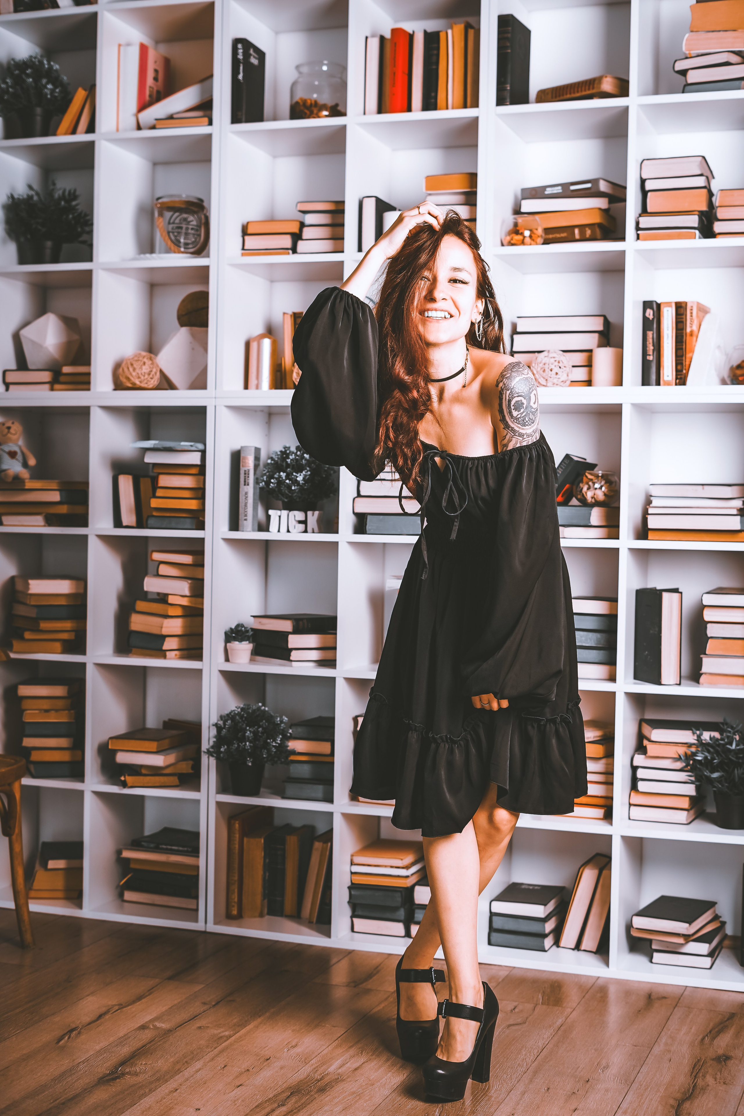 Девушка в красивом черном платье стоит у полки с книгами и смеется